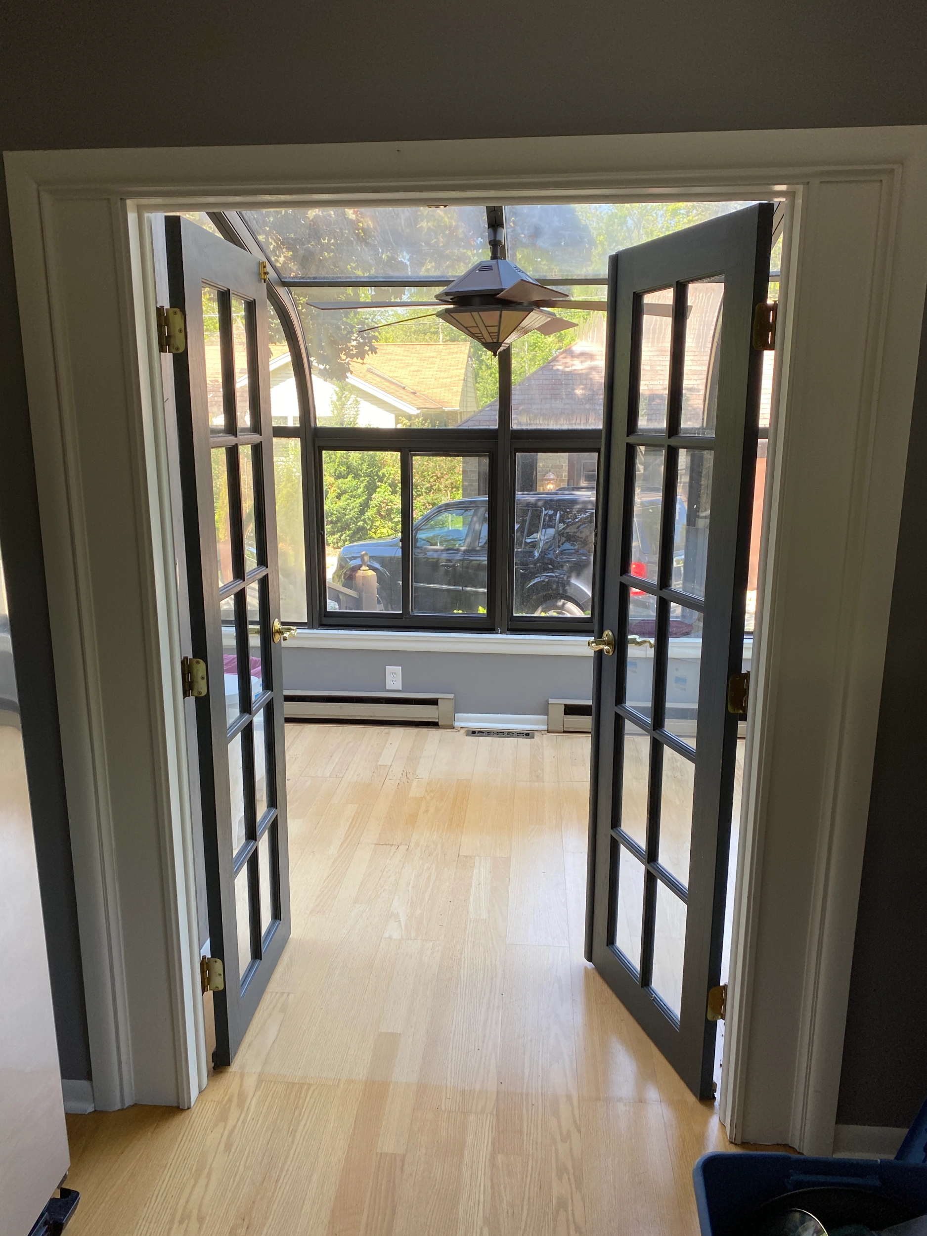 Open French doors lead to a sunlit room with wooden flooring. Outside, greenery, rooftops, and a parked vehicle are visible.