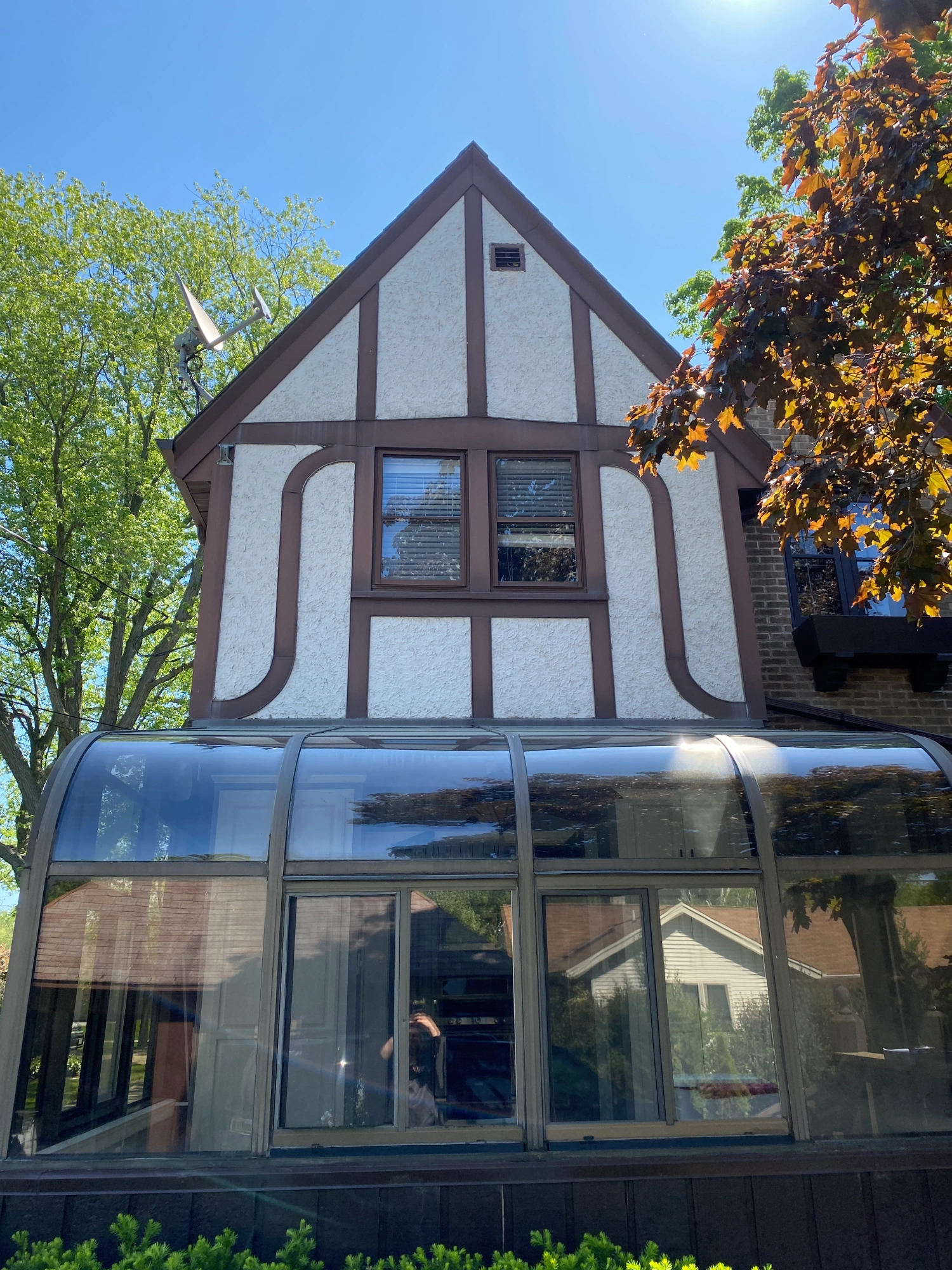 A Tudor-style house with wooden accents, large windows, and a glass sunroom. Branches frame the scene against a bright sky.