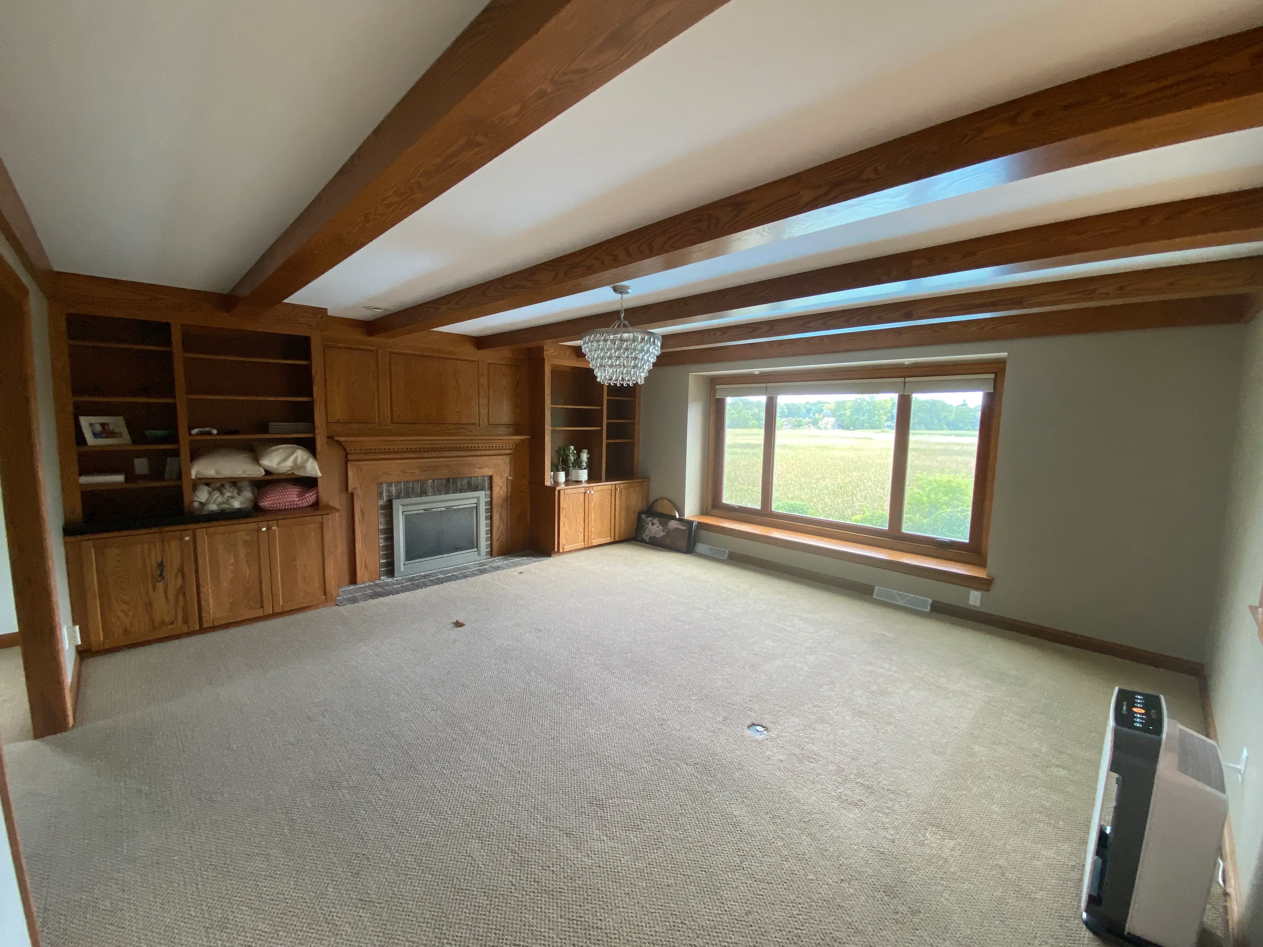 A cozy living room features wooden beams, built-in shelves, a fireplace, and a large window overlooking fields, with a decorative light fixture.