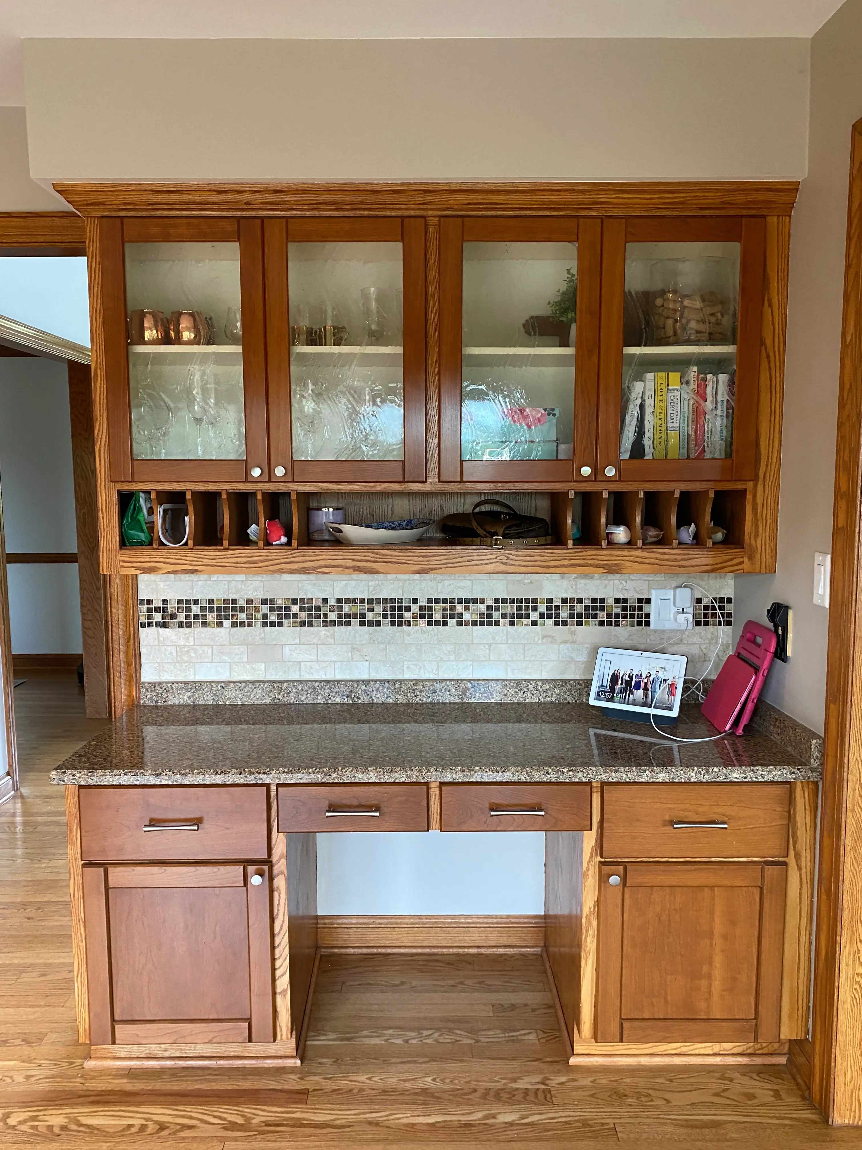 A wooden cabinet with glass doors, granite countertop, and a photo frame on display. Kitchen items and books are stored inside.