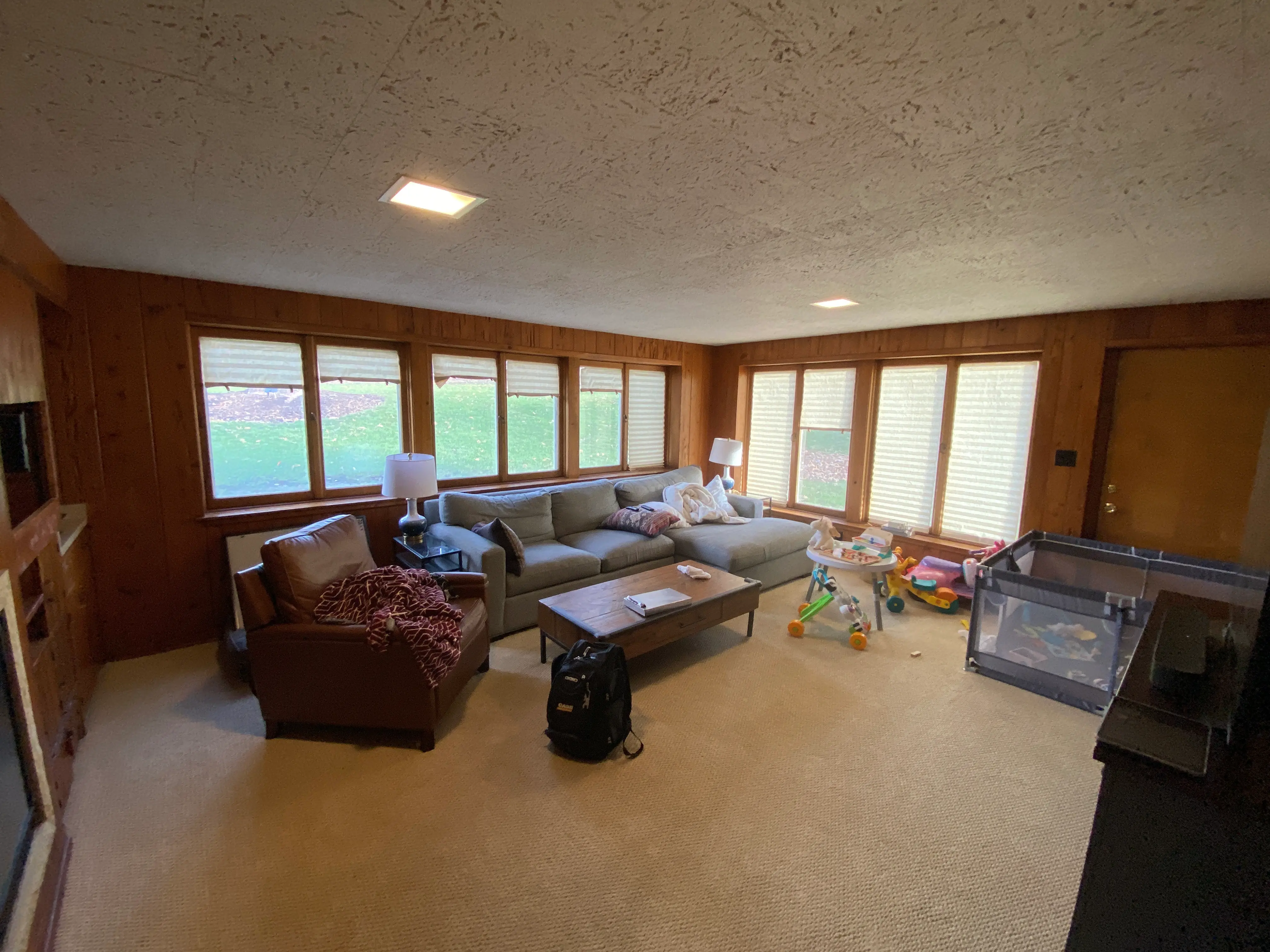 Cozy living room with large windows, sectional sofa, armchair, toys, and playpen. Natural light bathes the wooden-paneled space, creating a warm atmosphere.