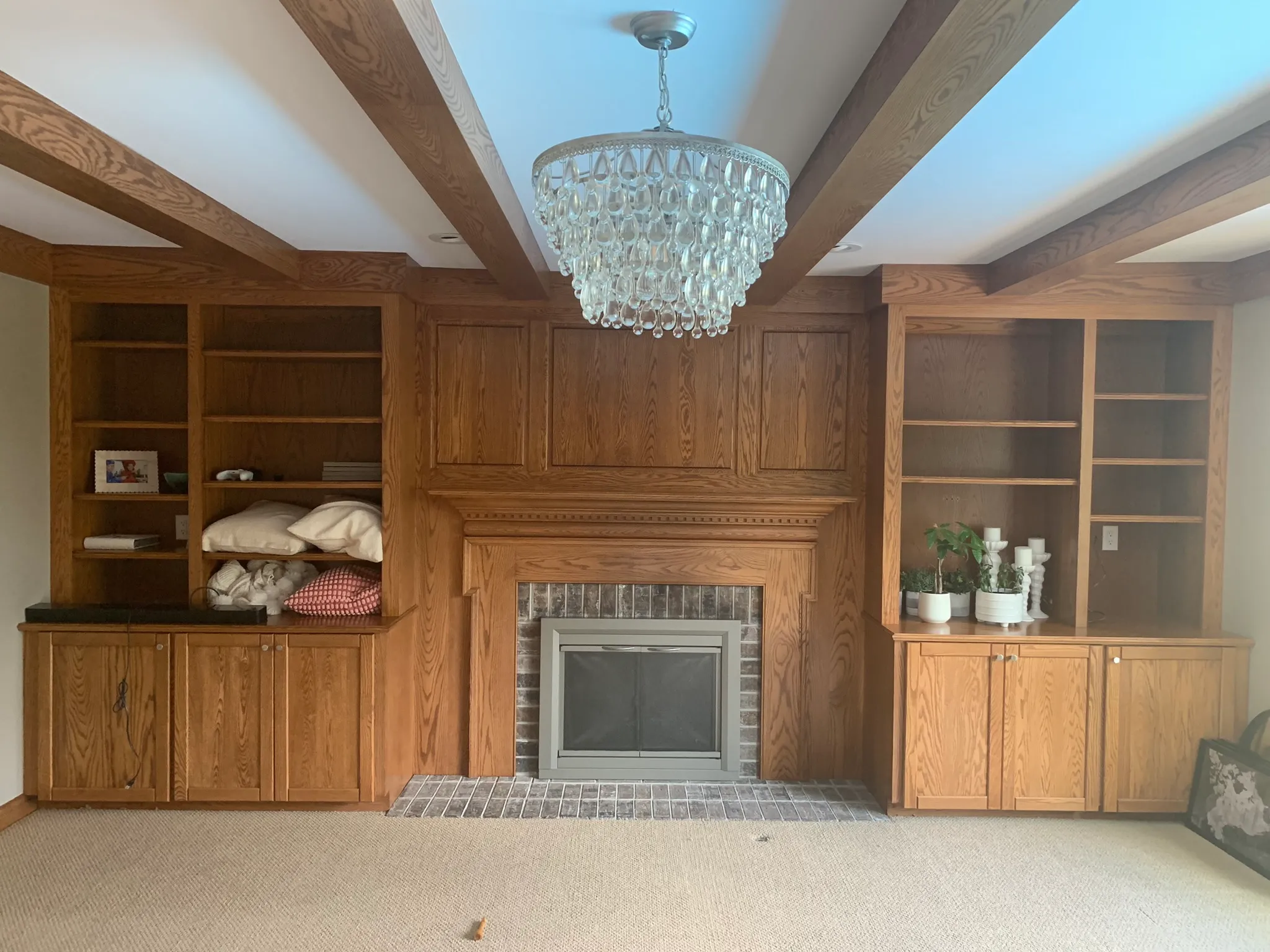 A cozy room features a wooden fireplace, built-in shelves, a crystal chandelier, and houseplants. Pillows and decor items add a homely feel.