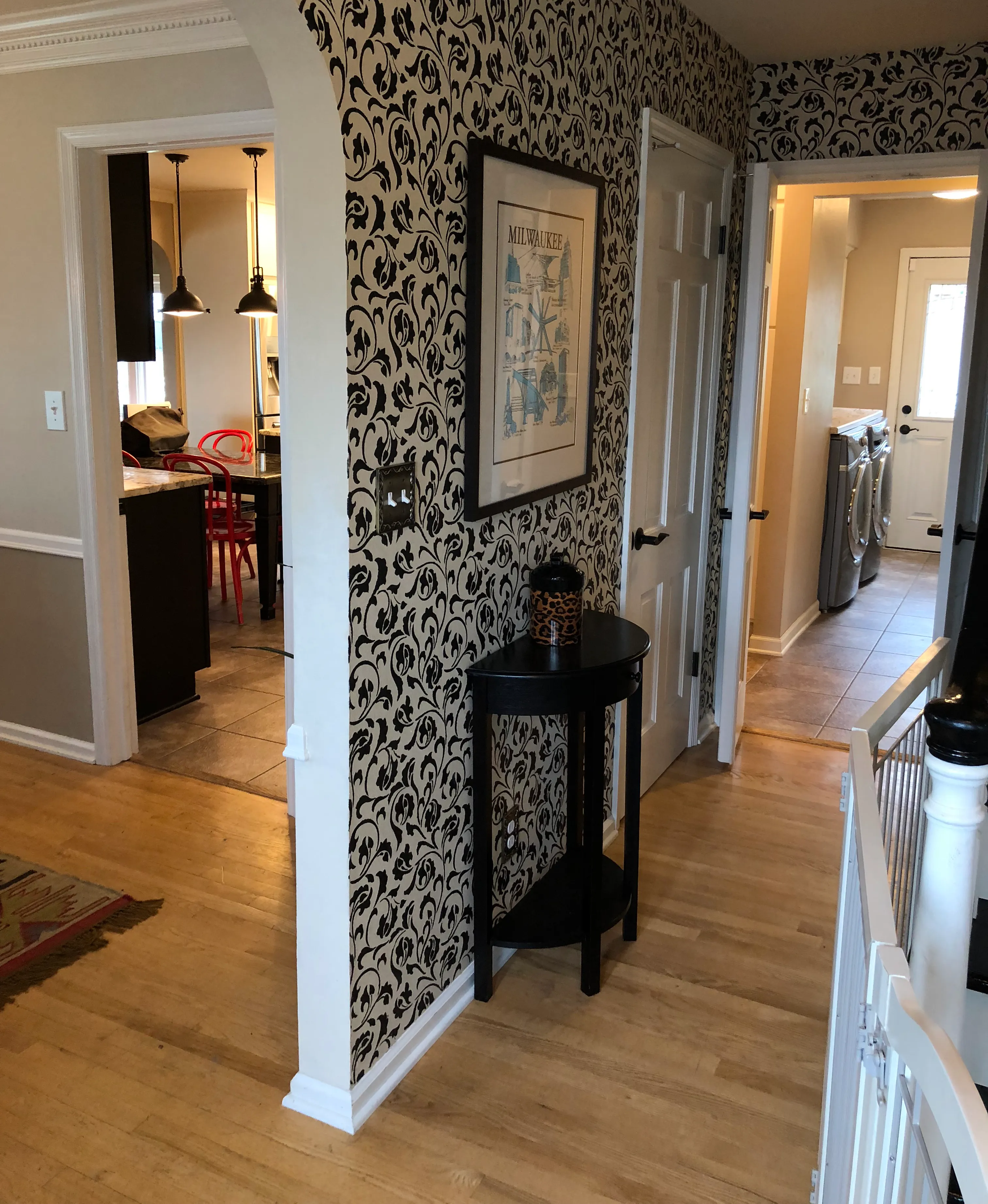 Decorated hallway featuring patterned wallpaper, framed Milwaukee map, wooden flooring, small black table, adjacent dining area with chairs, and laundry room in view.