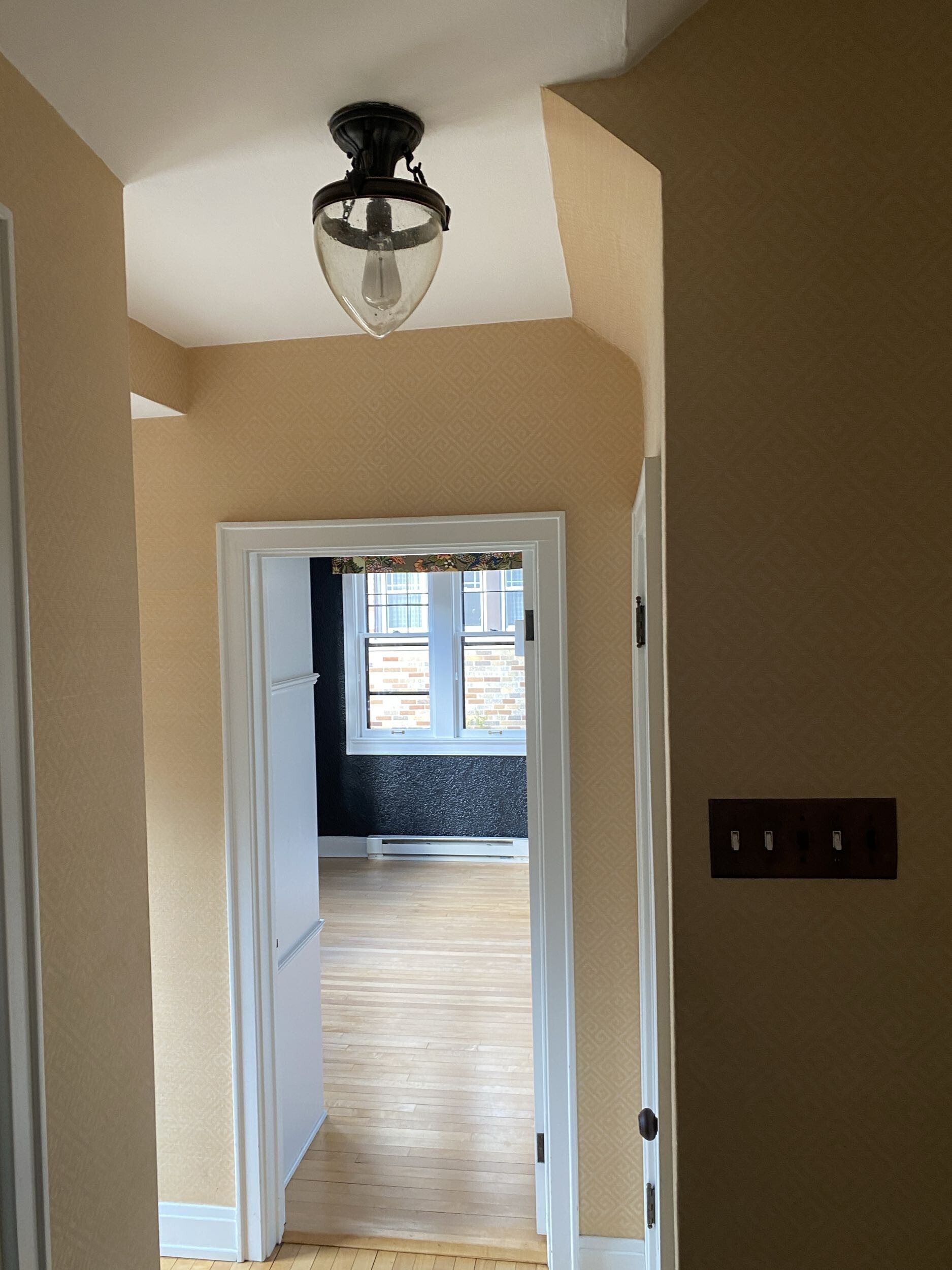 A hallway with beige walls leads to a bright room with wooden floors, featuring a vintage light fixture and visible windows.