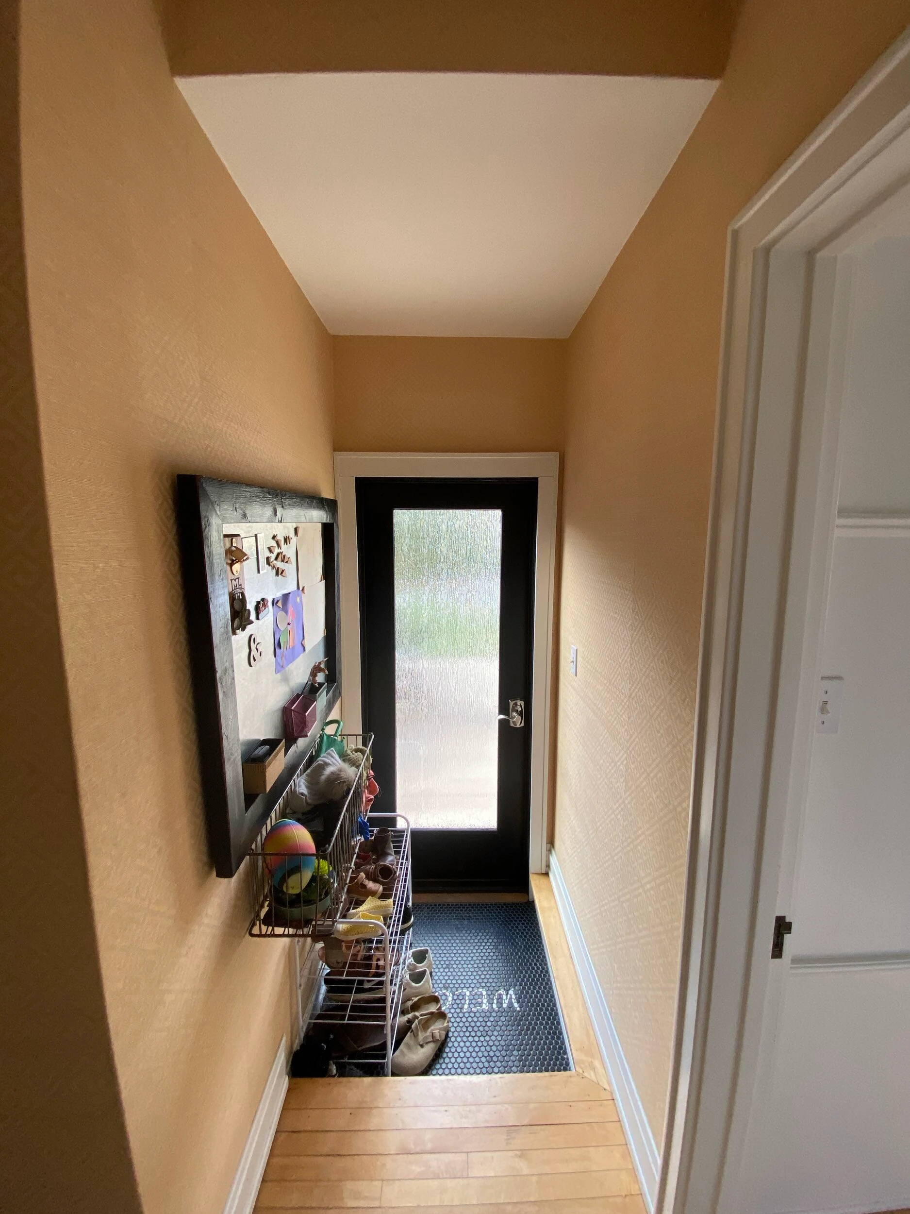 A narrow entryway with a welcome mat, shoe rack, and bulletin board beside a black-framed door with frosted glass.