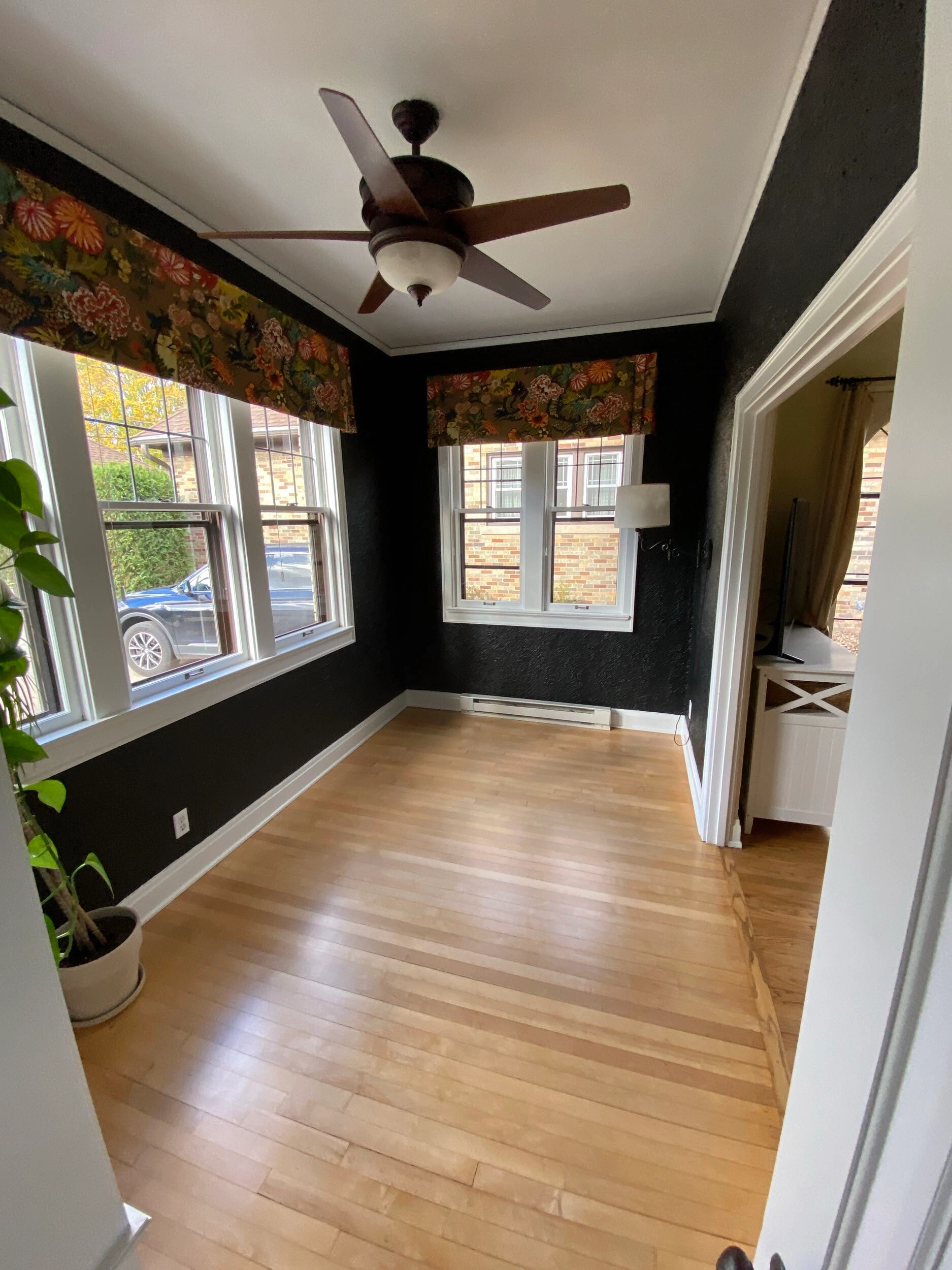 A bright, empty room with wooden floors, floral curtains, and a ceiling fan. Large windows provide natural light, revealing a car outside.