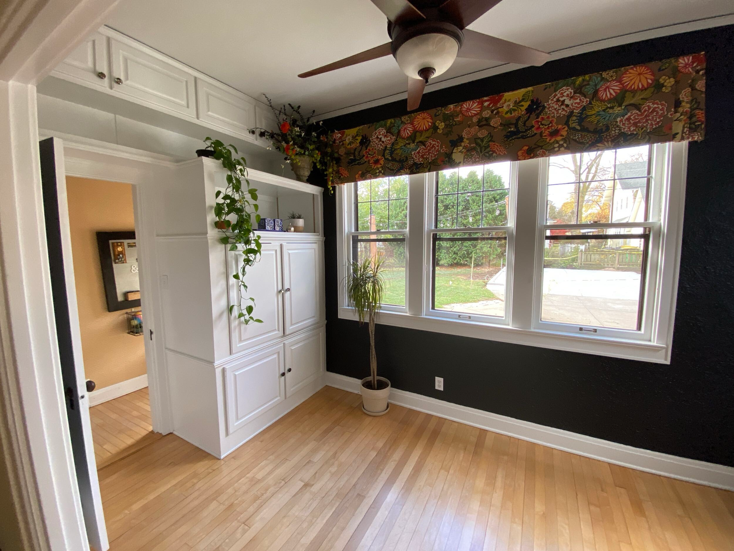 Room with hardwood floor, large windows, floral valance, ceiling fan, white cabinets, and indoor plants, overlooking a green garden and trees outside.