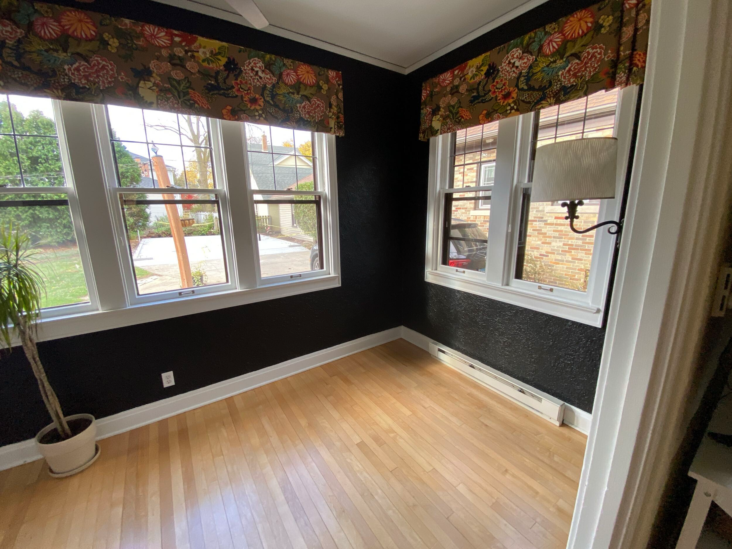 A sunlit room with large windows, floral curtains, light wood flooring, a potted plant, and a wall lamp, overlooks a suburban street.