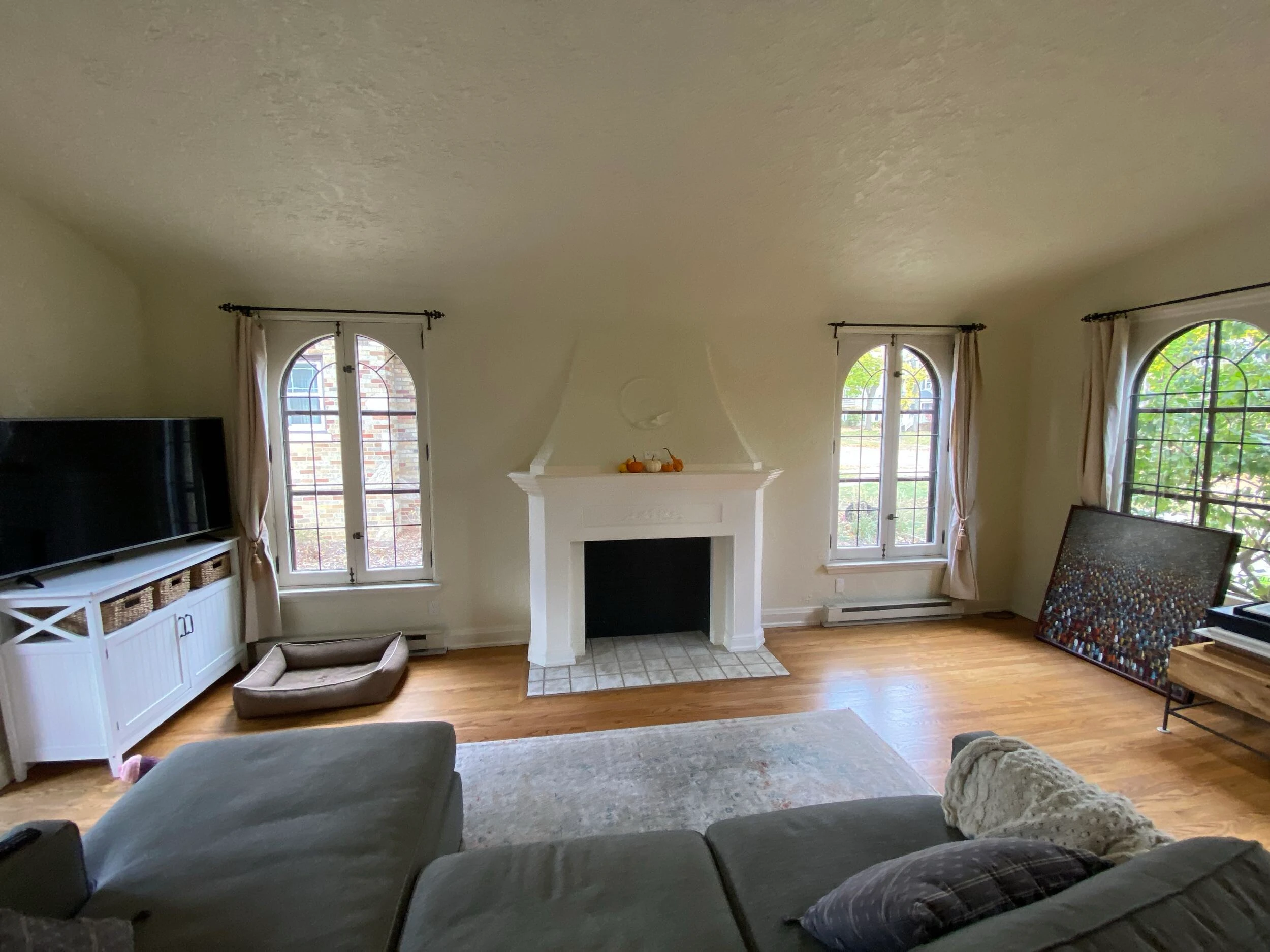 A cozy living room with arched windows, a white fireplace, large TV, pet bed, and a painting on the floor.