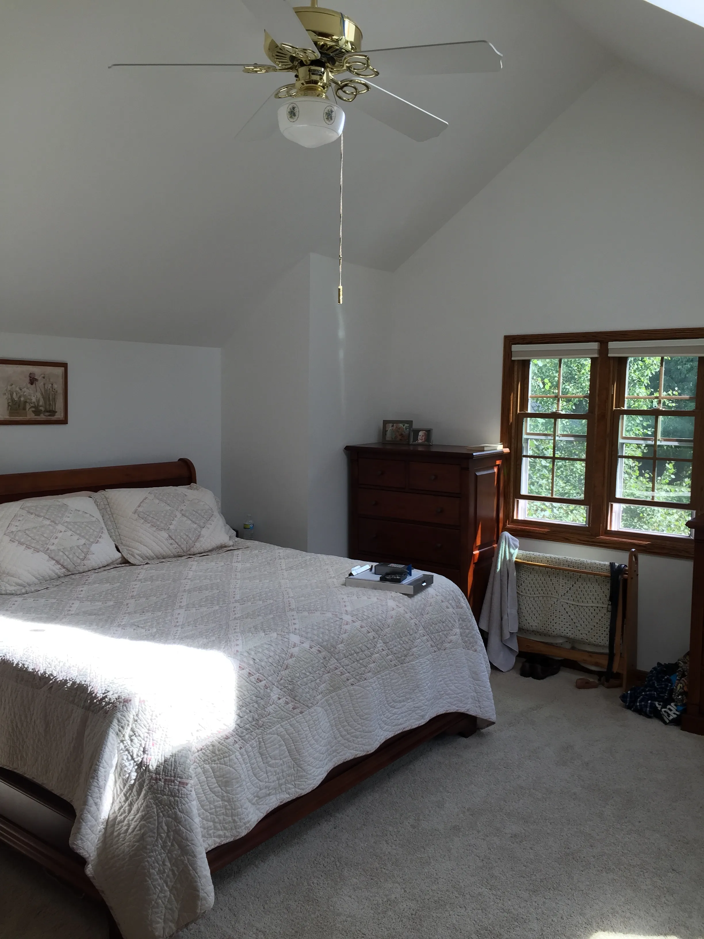 A cozy bedroom with a quilted bed, wooden furniture, and a ceiling fan. Sunlight streams through the window, brightening the space.