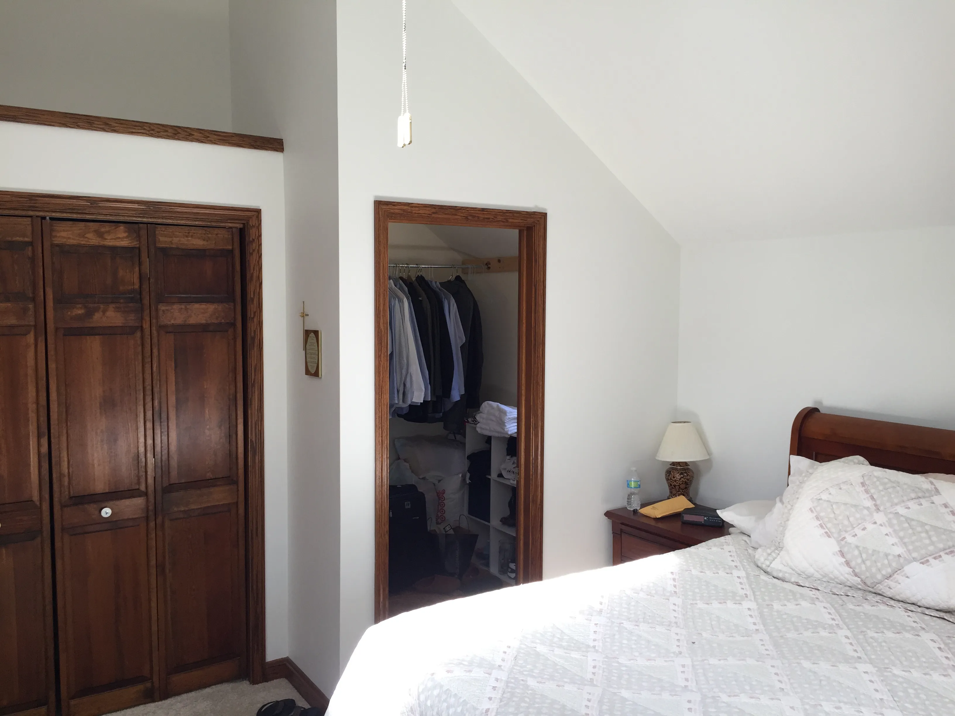 A cozy bedroom with a wooden closet, open door to a closet with hanging clothes, and a bed beside a small lamp on a nightstand.