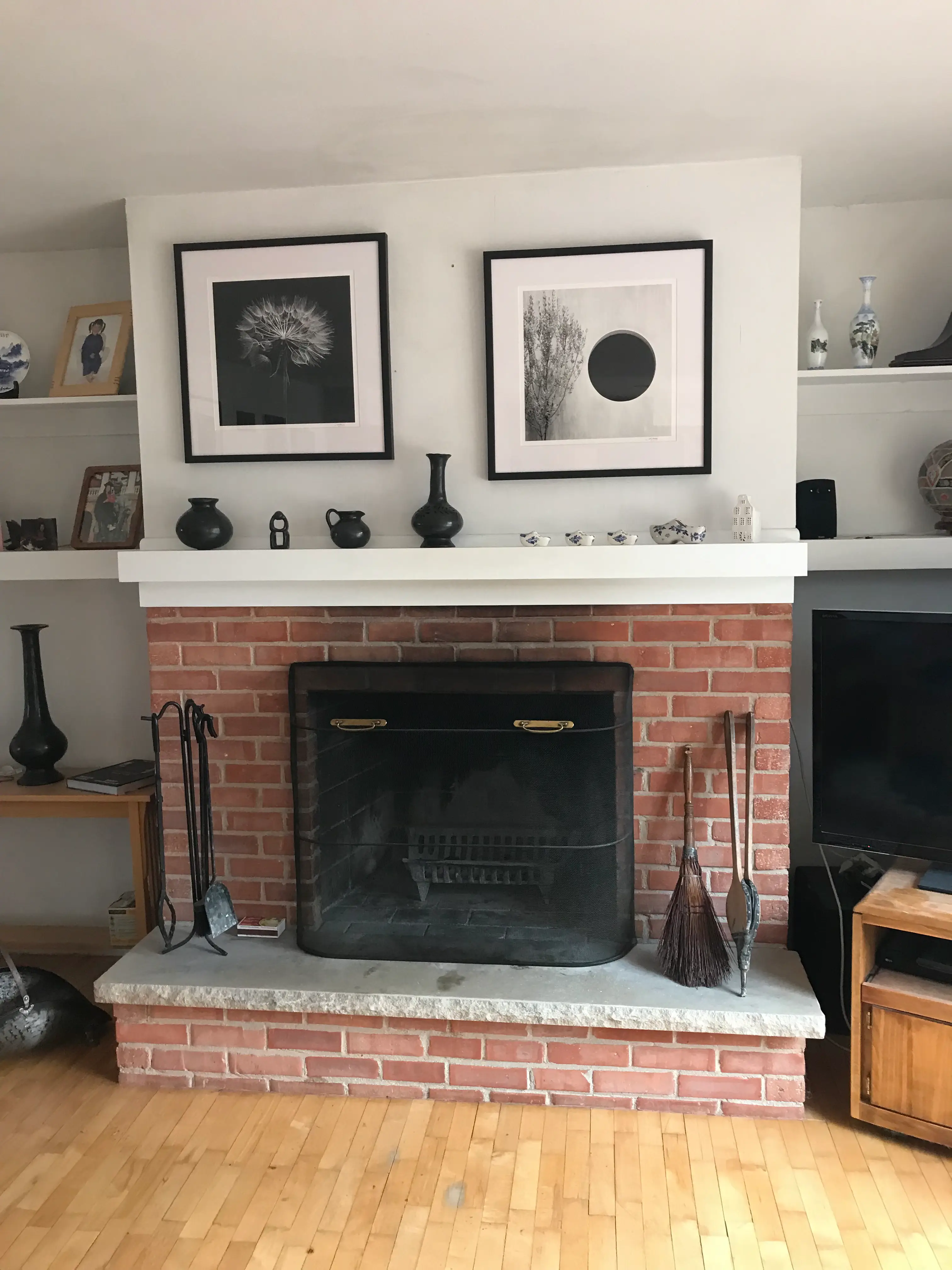 A cozy living room features a brick fireplace with framed artwork above, surrounded by decorative vases on shelves and black fireplace tools.