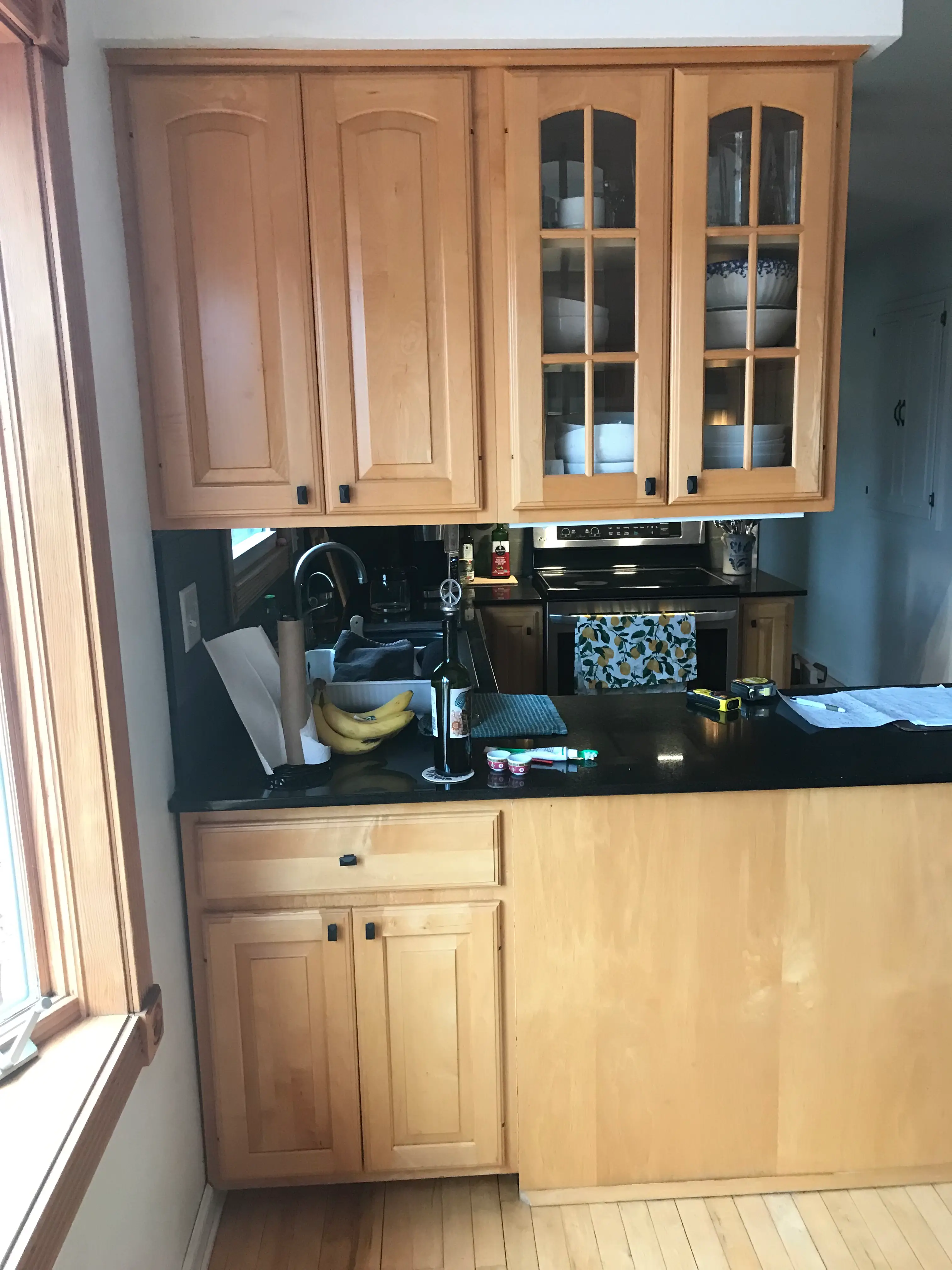 A kitchen features wooden cabinets, a black countertop with bananas, wine, and papers. A window provides natural light, creating a cozy atmosphere.