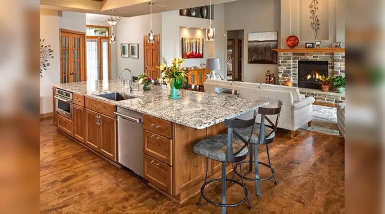 Modern kitchen with granite island, wooden cabinets, and cozy living area featuring a fireplace. Bright, open space with stylish decor and plants.