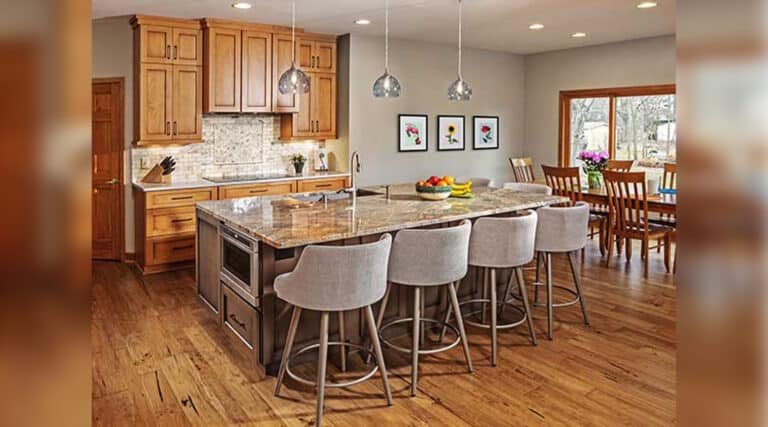 Modern kitchen with wooden cabinets, granite island, and bar chairs. Dining table with flowers is near a large window. Artwork decorates the walls.