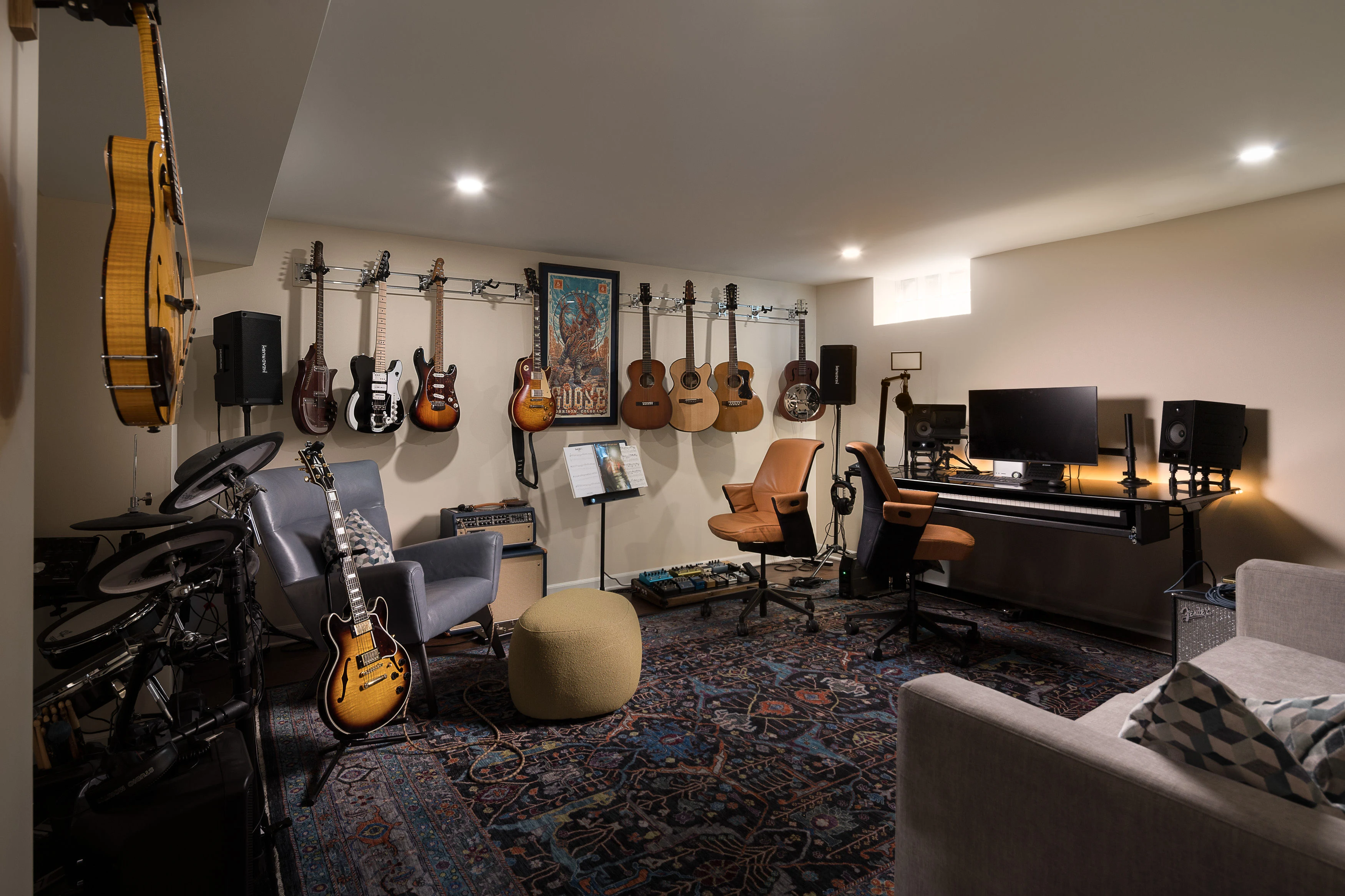 A music studio features hanging guitars, a drum set, computer workstation, chairs, and vibrant rug, creating a cozy and creative recording environment.