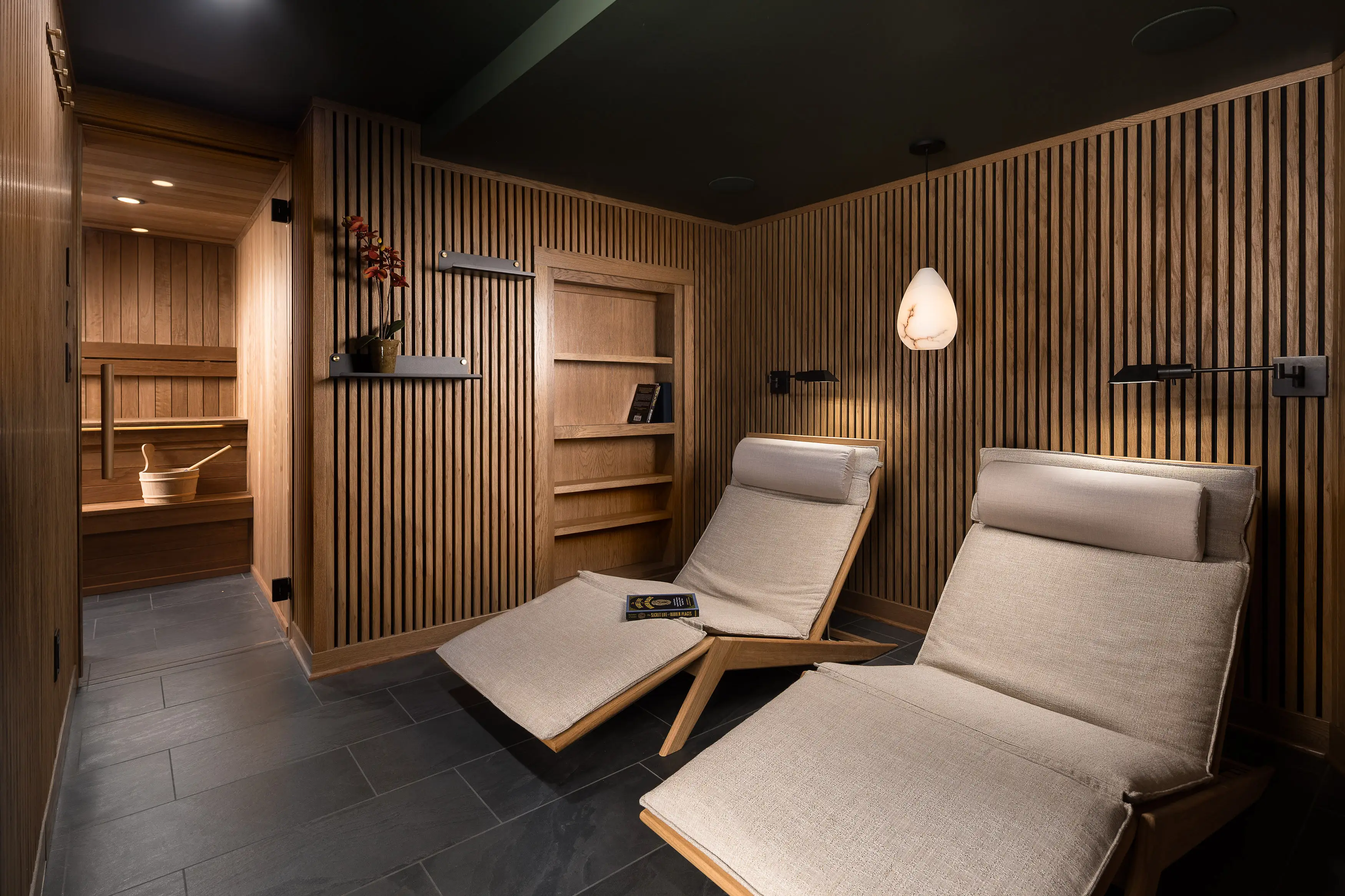 A cozy, modern relaxation room with wooden lounge chairs, wall paneling, a pendant light, and an adjoining sauna featuring a wooden bucket.