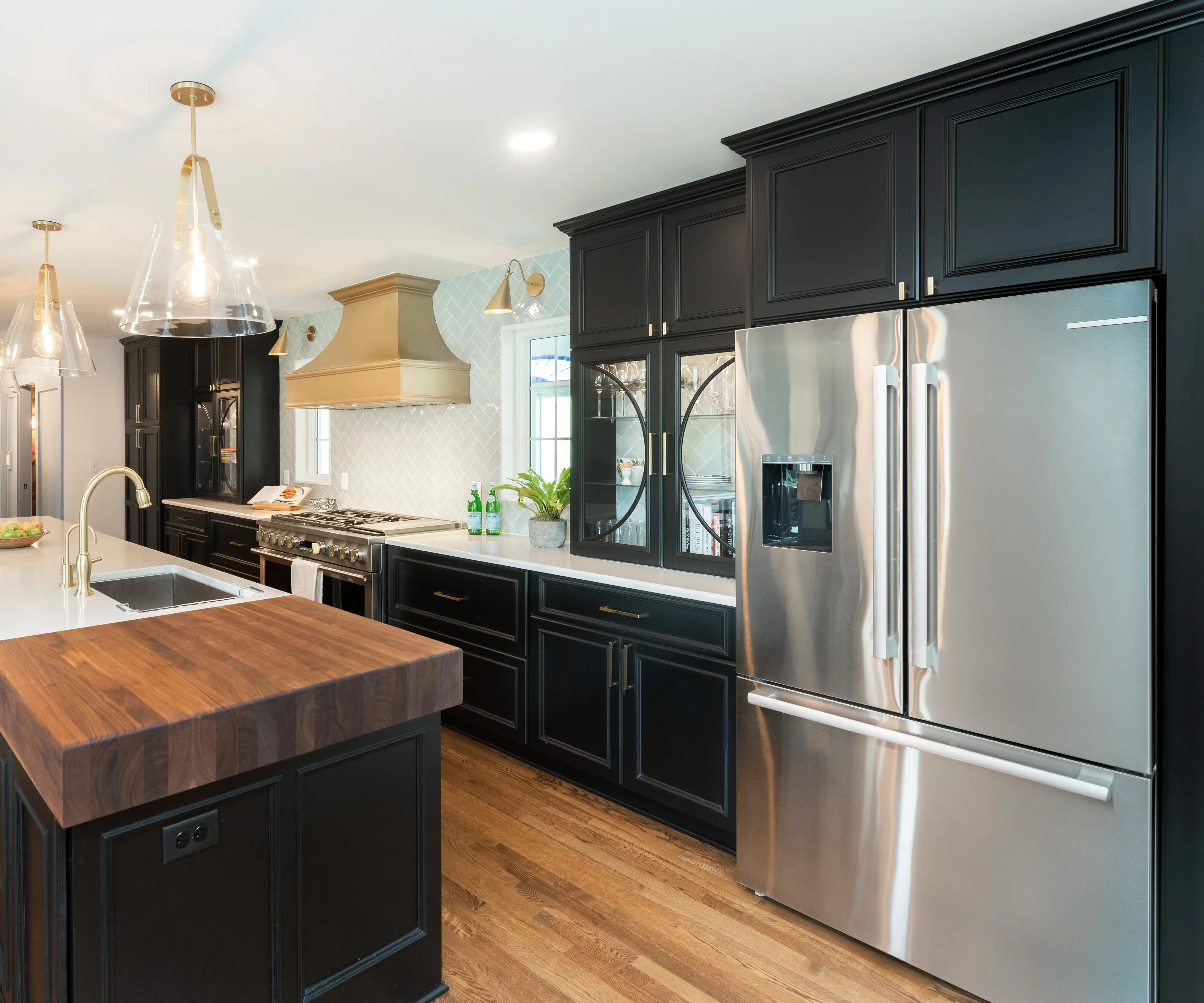 Modern kitchen with black cabinetry, stainless steel appliances, wooden island, and pendant lights. Features a stylish hood and plant decoration. Wood flooring.