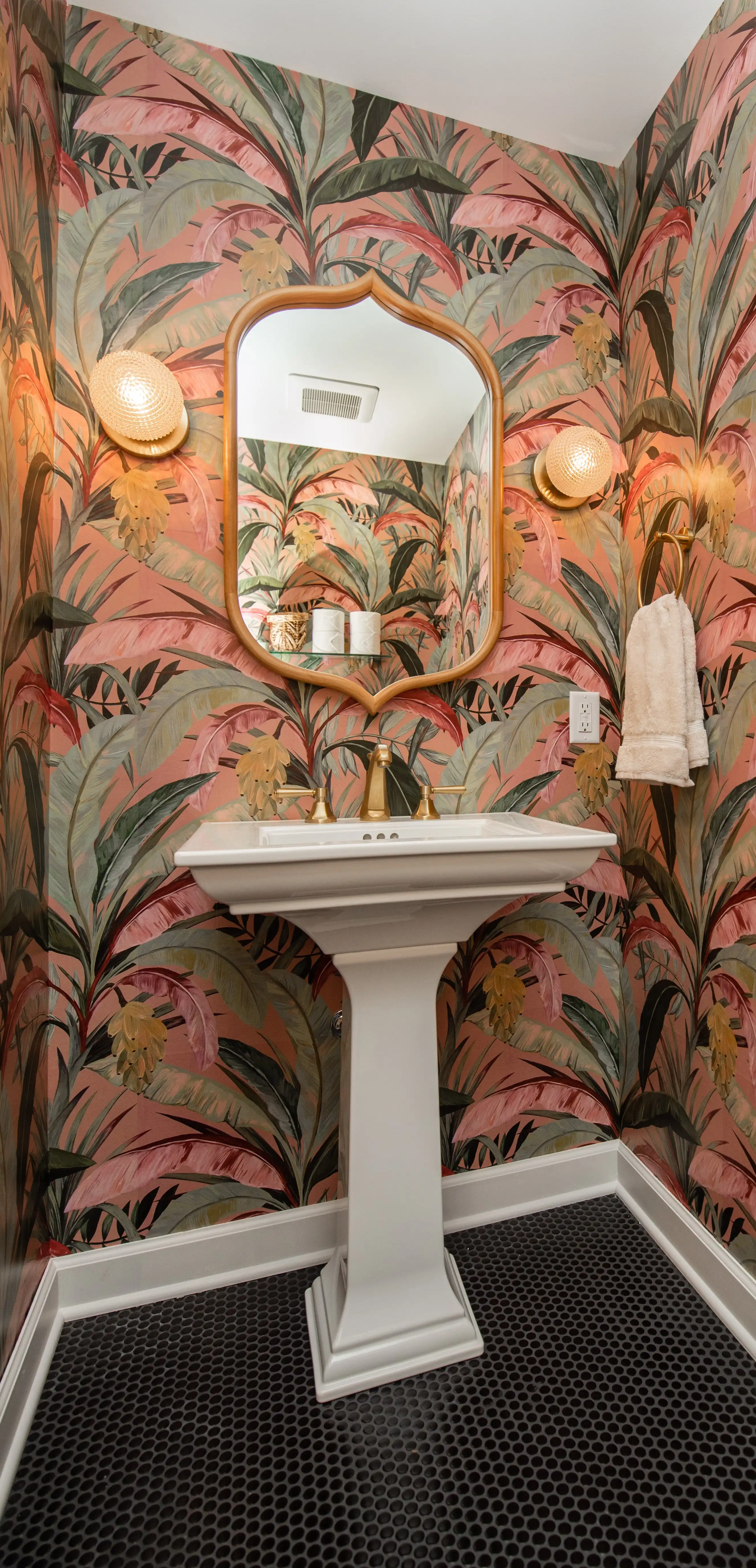 A bathroom with vibrant tropical wallpaper, pedestal sink, decorative mirror, towel rack, and globe sconces creates a warm, inviting atmosphere.