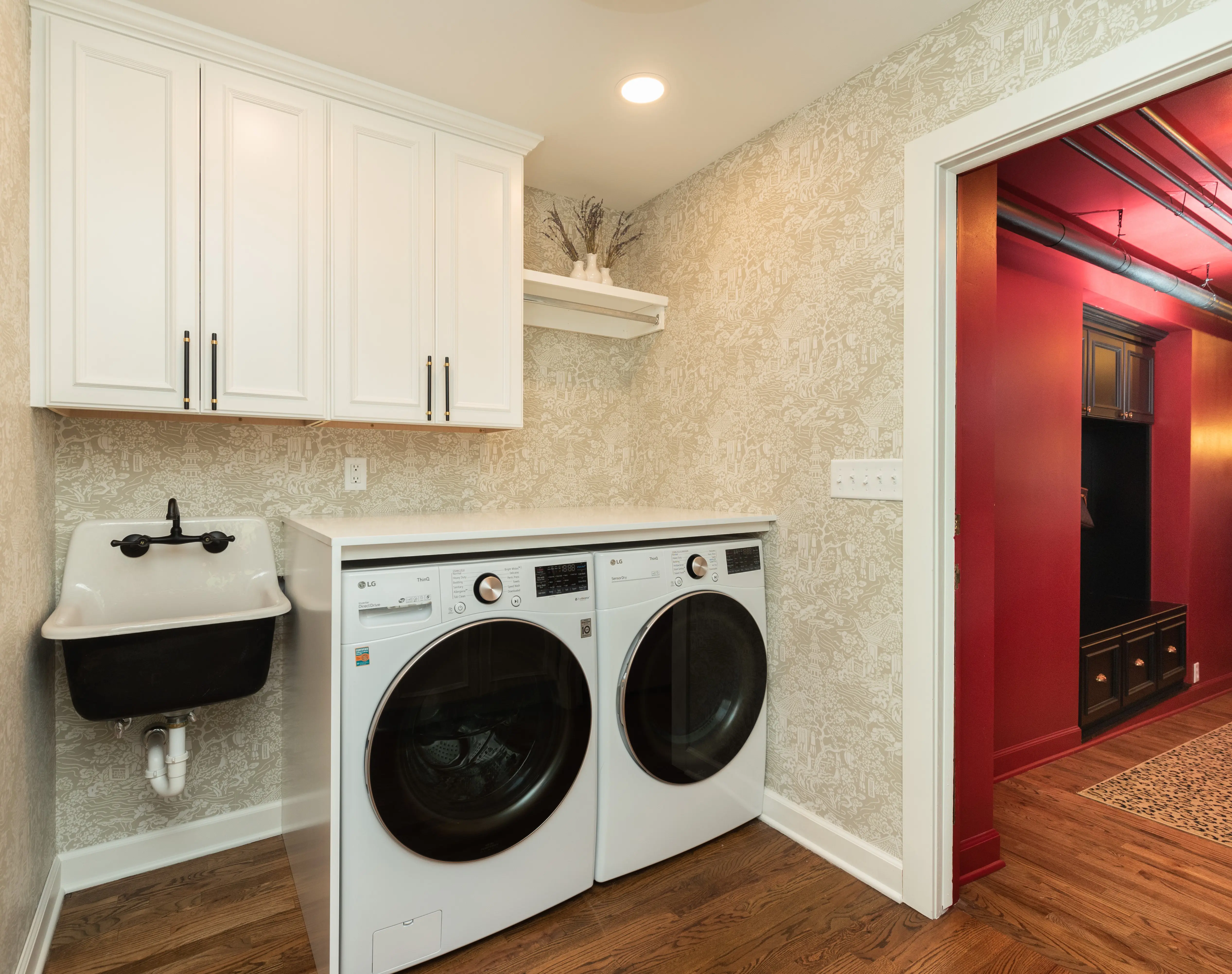 A modern laundry room features a washing machine, dryer, white cabinets, black sink, patterned wallpaper, and a doorway leading to a vibrant red room.