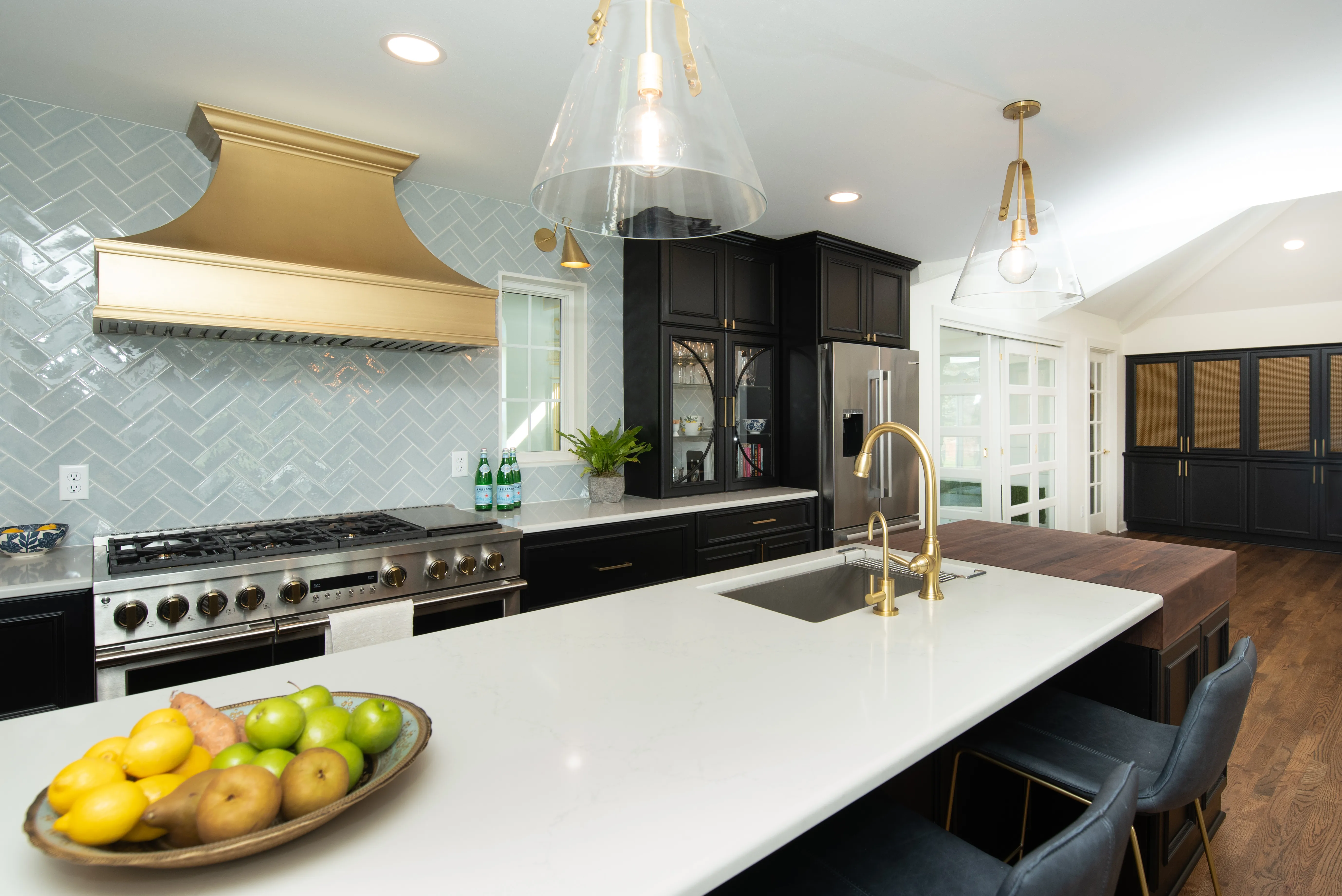 Modern kitchen with stainless steel appliances, black cabinets, gold accents, and a marble island. Bowl of fruit adds a fresh touch.