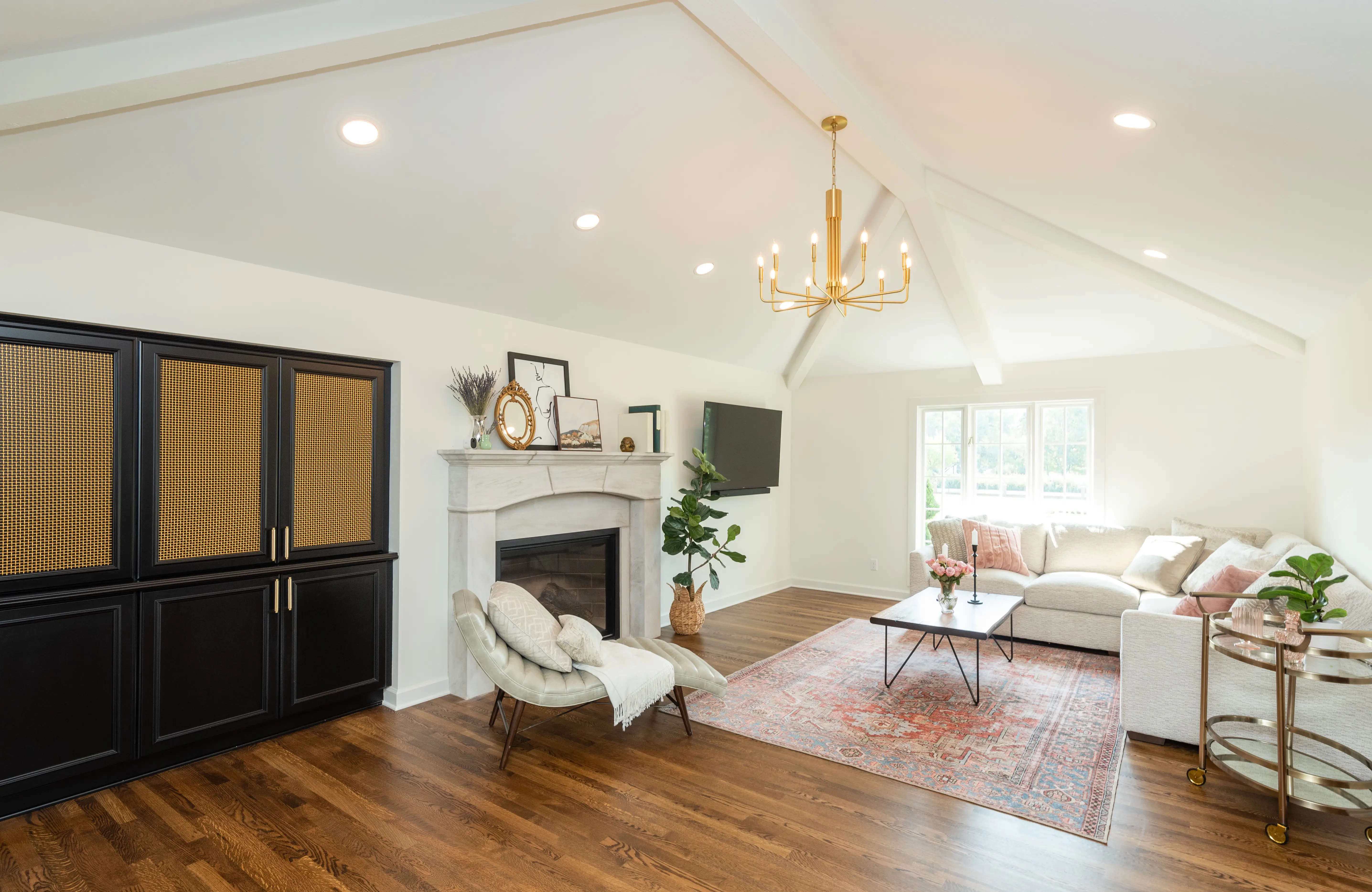 Bright living room with vaulted ceiling, chandelier, fireplace, cozy seating, and large windows. Wooden floor complements elegant decor and stylish furniture arrangement.