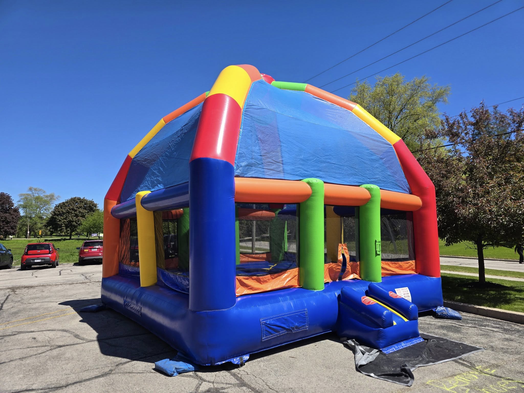 A colorful inflatable bounce house is set up in a parking lot, surrounded by parked cars and green trees under a clear blue sky.