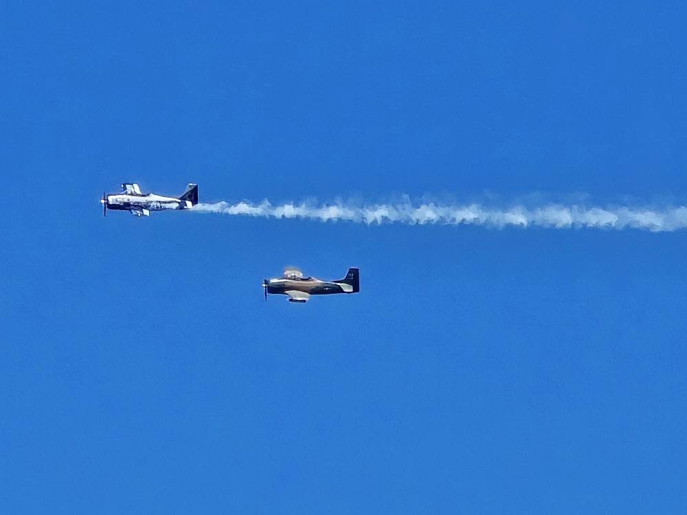 Two vintage airplanes fly in clear blue sky, emitting white smoke trails, showcasing aerial precision and synchronized flight.