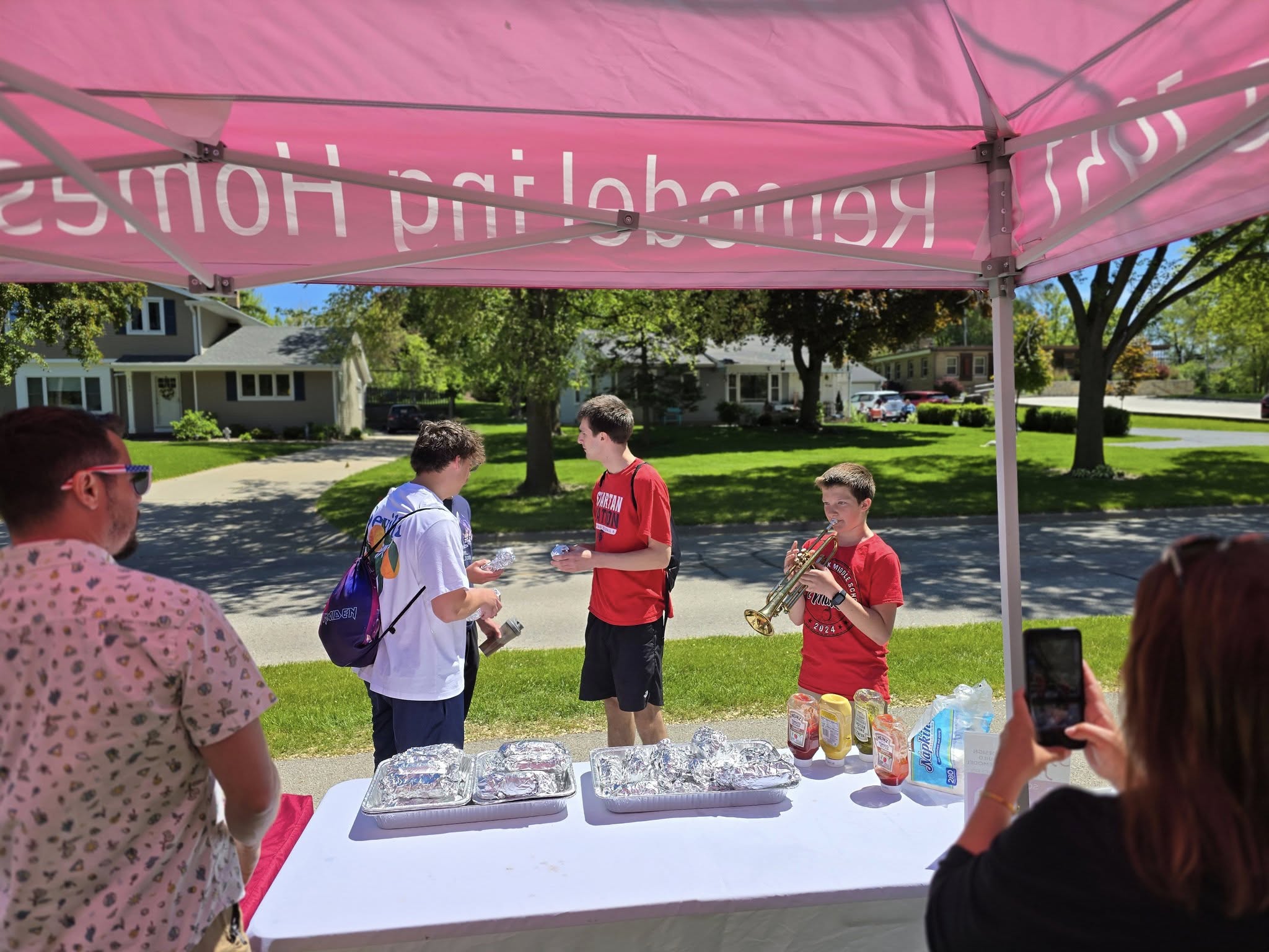 A group under a pink tent holds a casual outdoor event with food while a person plays trumpet in a suburban neighborhood setting.