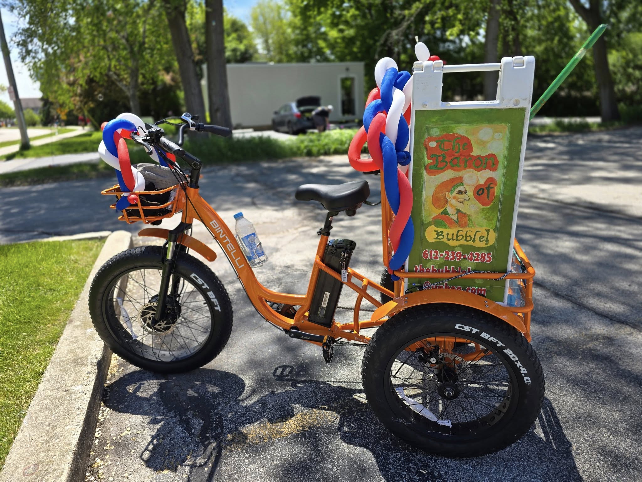 An orange tricycle with a cooler labeled "The Baron of Bubble" on a sunny street, decorated with red, white, and blue balloons.