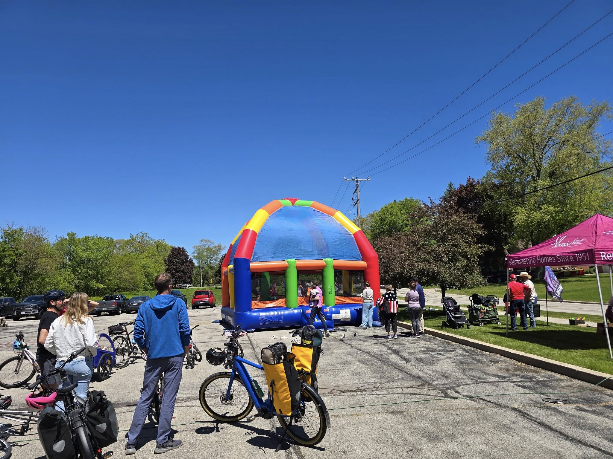 People gather around bikes and a colorful inflatable bounce house, with a pink tent nearby, under a clear blue sky.