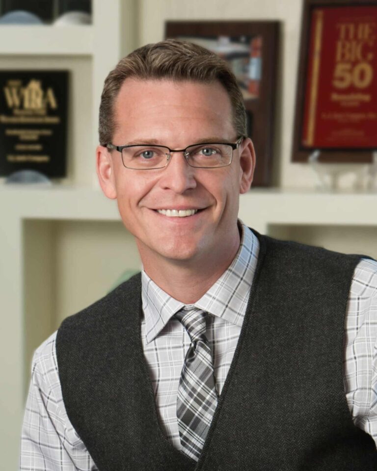 A person wearing glasses and a checked shirt, smiling in an office setting with award plaques displayed in the background.