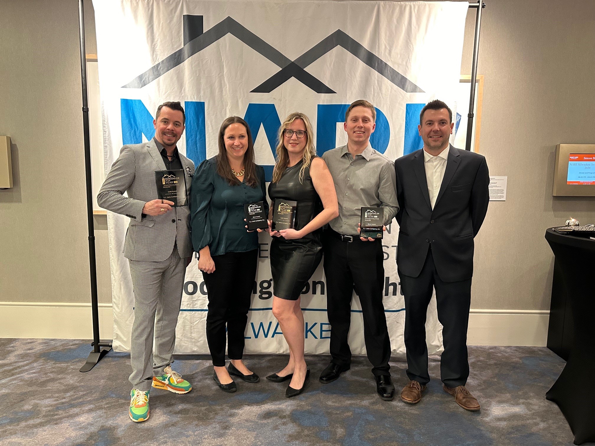 Five people pose with awards in front of a banner. The event appears professional, with individuals dressed in formal and semi-formal attire.