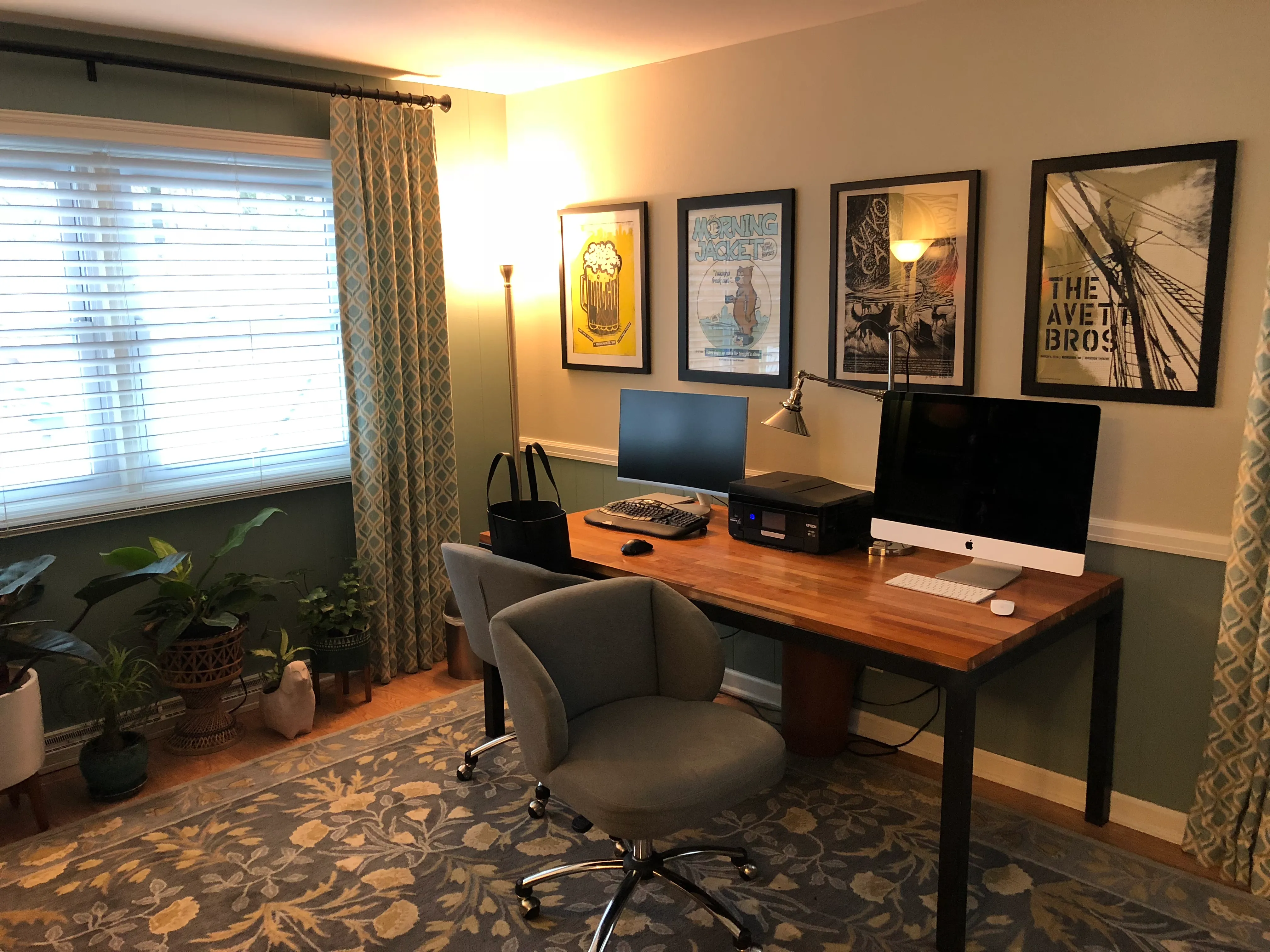 A home office features a wooden desk with computer, task lamp, and printer. Artwork and plants decorate the space, enhancing its cozy ambiance.
