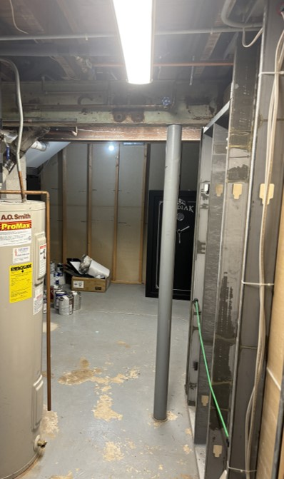 A dimly lit basement with a water heater, pipes, exposed beams, and cluttered items in the corner, creating an unfinished, utilitarian space.