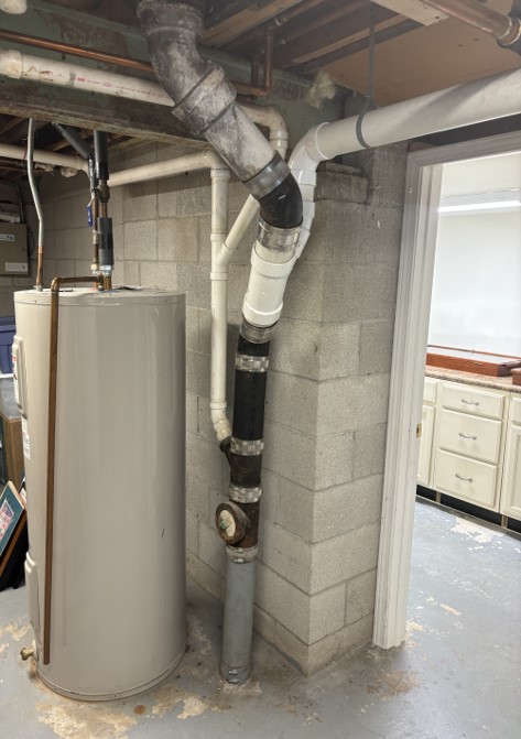 A basement with exposed pipes and a hot water heater against a cinder block wall. Adjacent room with cabinets and countertop visible.