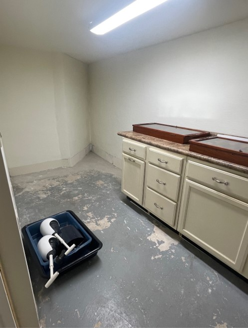 A small, undecorated room with bare walls. There is a cabinet with drawers, and plastic containers holding dome-shaped objects on the floor.