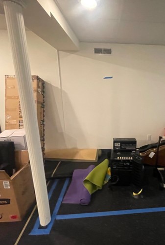 A basement room with stacked boxes, a support column, rolled yoga mats, and gym equipment on the floor. Walls are painted white.