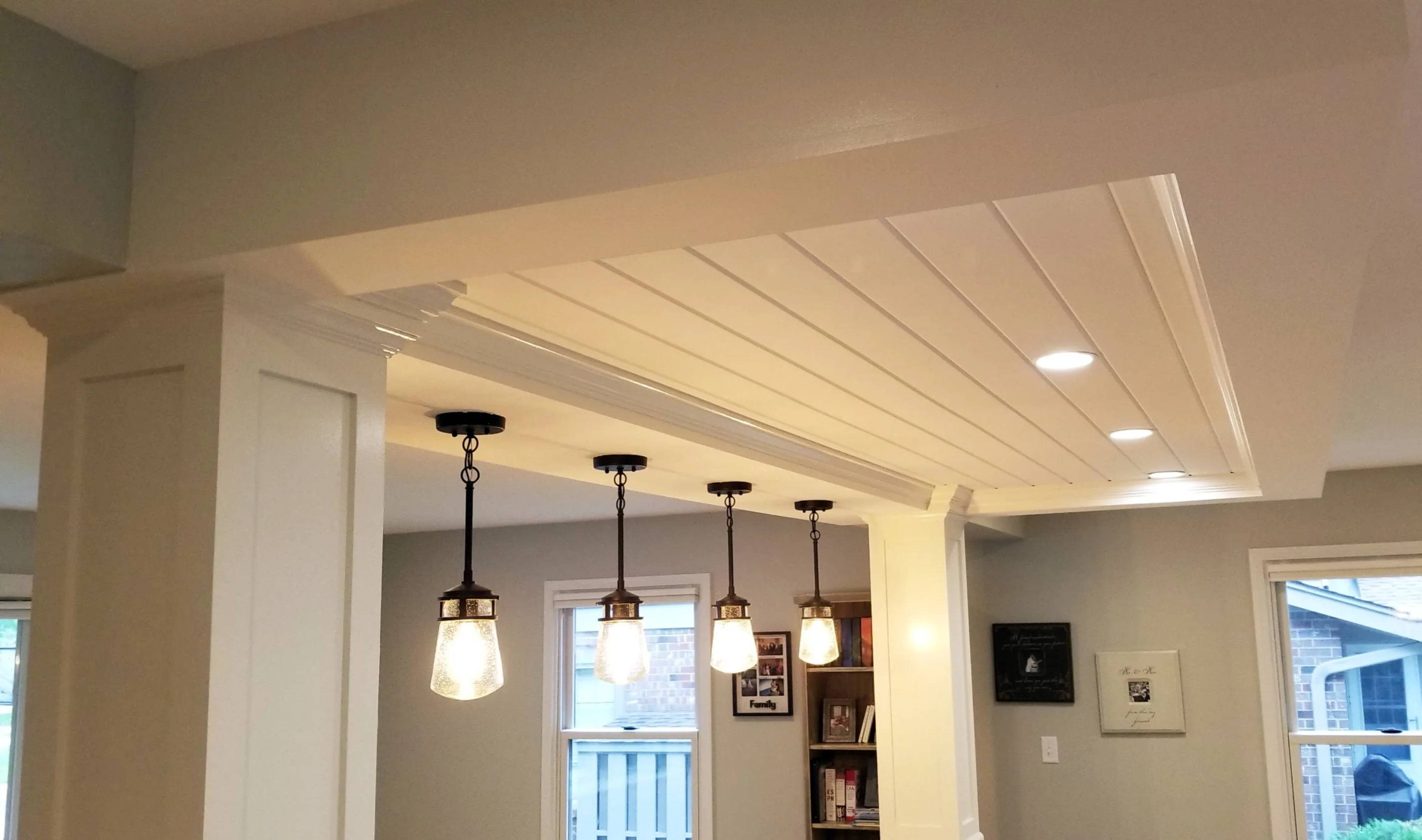 Modern kitchen ceiling with exposed beams, hanging lights, recessed lighting, and adjacent wall shelves. Window view of brick building outside.
