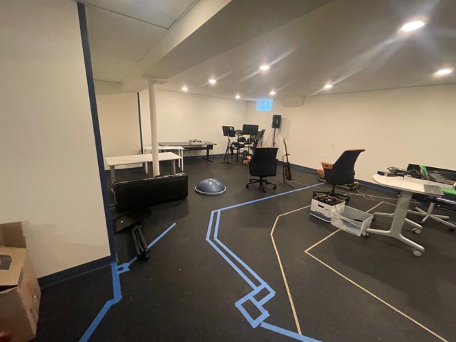 Spacious room with scattered chairs, tables, and musical instruments. Bright overhead lighting and blue tape patterns on floor, suggesting organized activities or workspace layout.