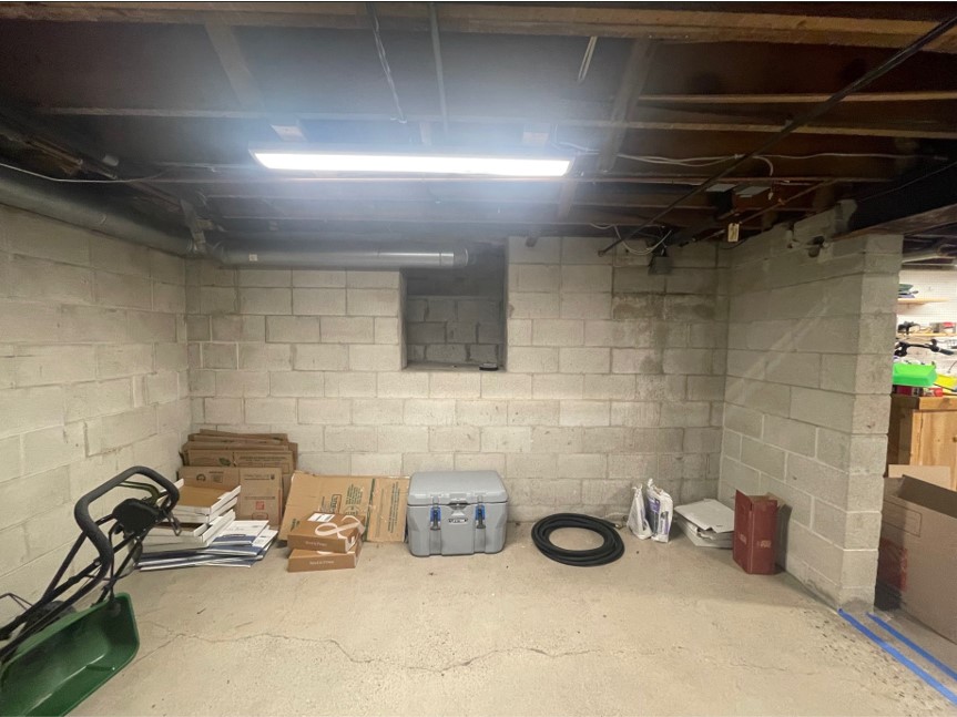 A basement storage area with concrete walls, various boxes, a cooler, and some equipment. Exposed ceiling beams and fluorescent lighting above.