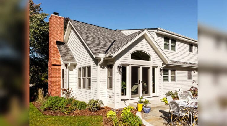 White house with brick chimney and gable roof. A patio with metal furniture is surrounded by plants. Clear sky overhead, creating a bright atmosphere.