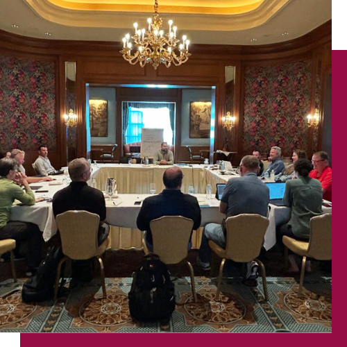 Meeting in an ornate conference room with a chandelier, featuring several people around a table with laptops and papers.