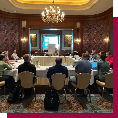 A group of people in a well-decorated room engaged in a meeting, seated around a table with laptops and papers.