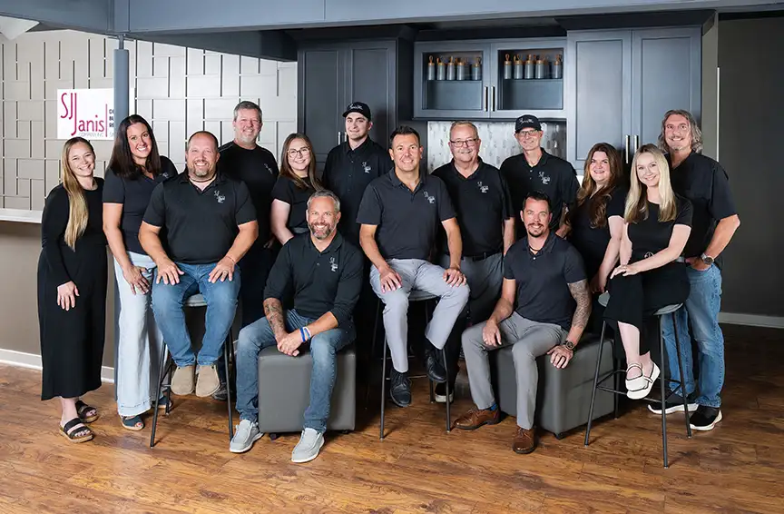 A group of sixteen people indoors, sitting and standing, casually dressed, smiling for a group photo with a "SJI" logo in background.