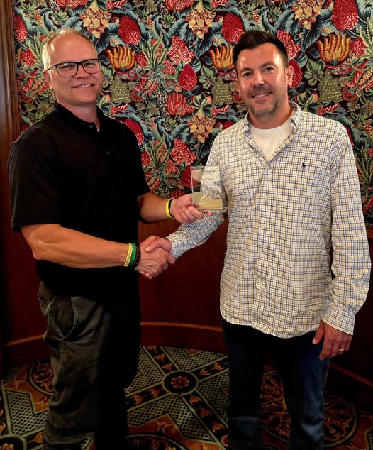 Two people shaking hands, one holding a clear plaque. Ornate floral wallpaper in the background. Patterned carpet underfoot.