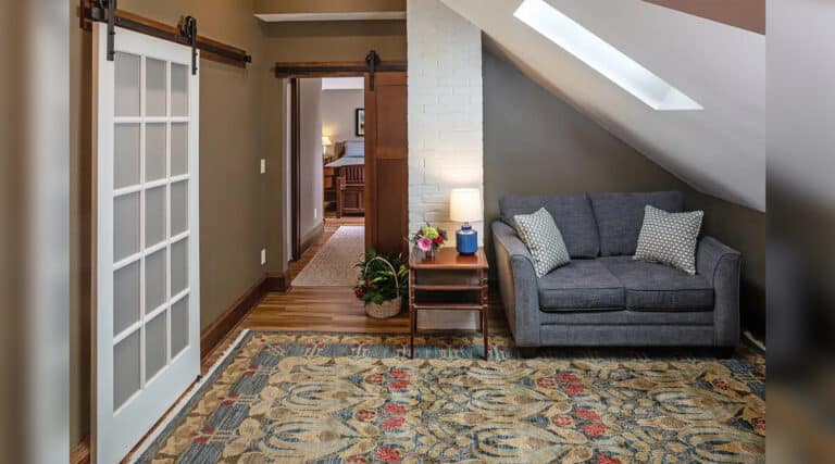 Cozy room with a gray sofa, patterned rug, sliding barn doors, and a side table holding a lamp and flowers. A hallway leads elsewhere.