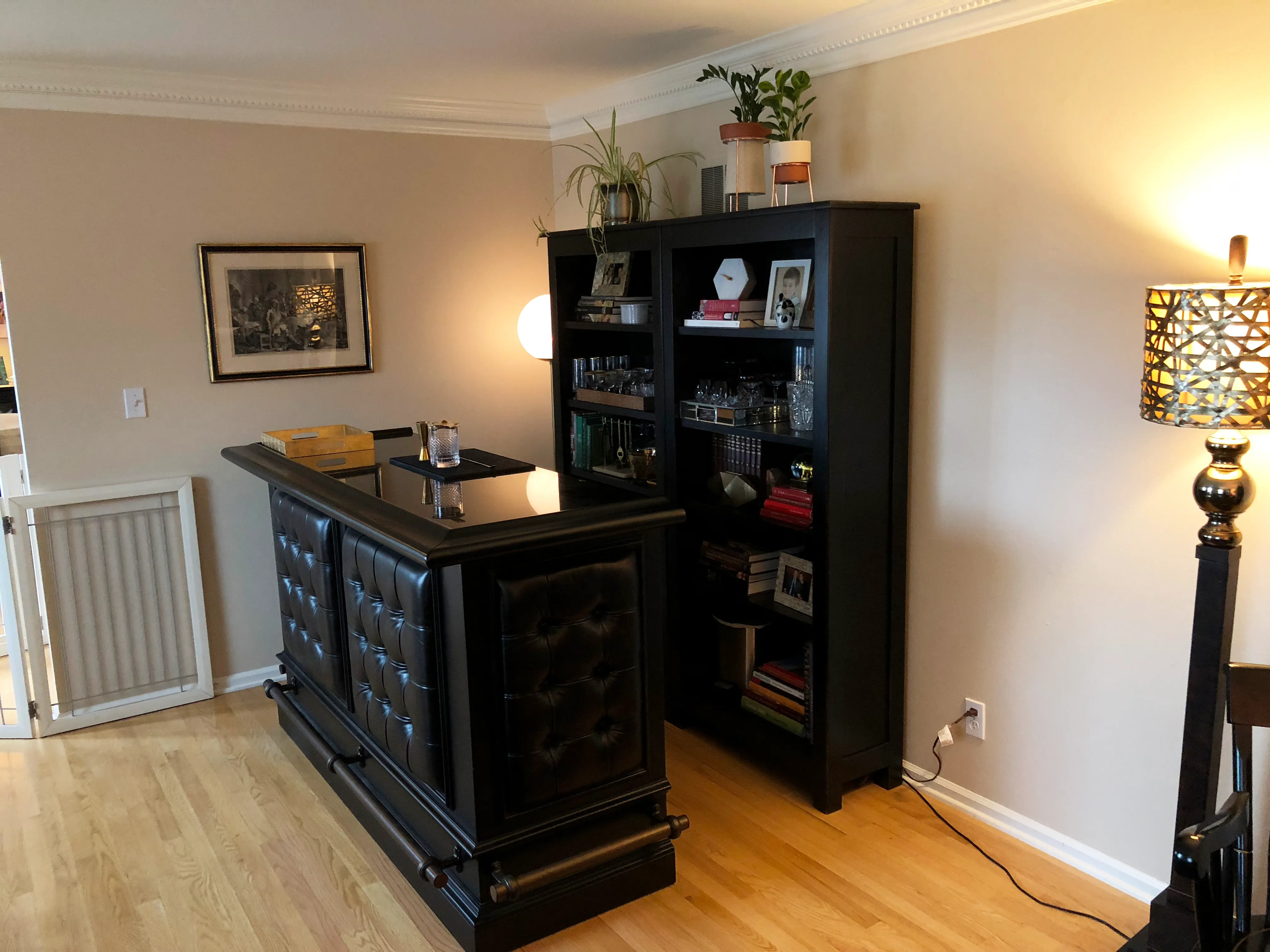 A home bar with black furniture, framed artwork, bookshelves, and potted plants on a wooden floor with ambient lighting.