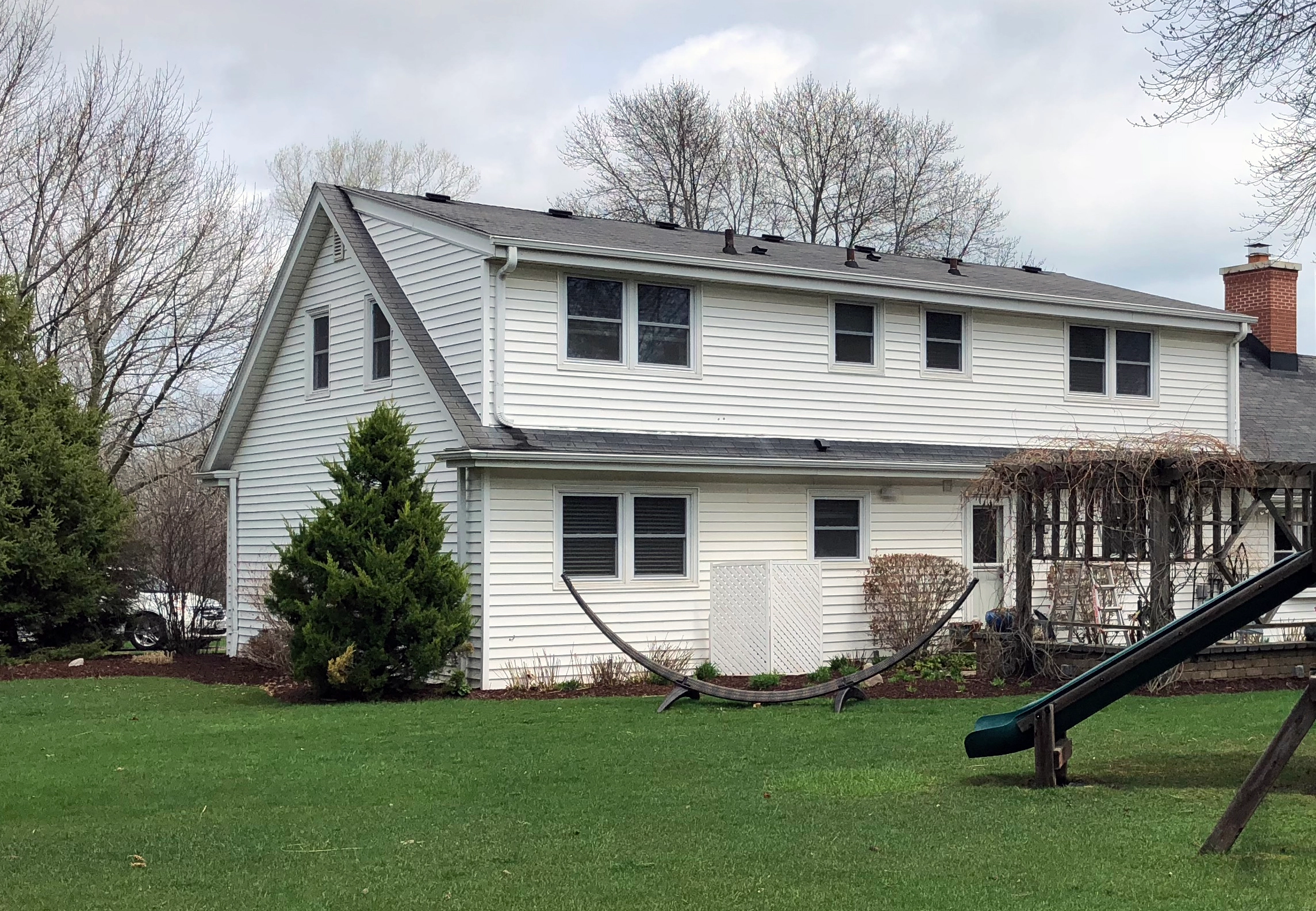 A two-story suburban house with white siding, surrounded by trees and a green lawn. Features a hammock stand and wooden slide.