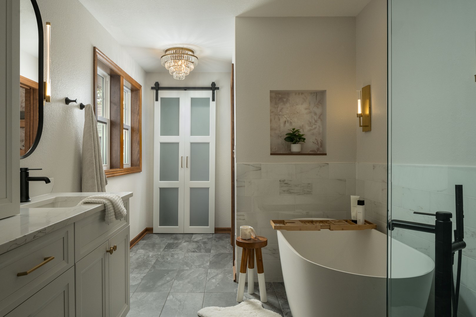 Elegant bathroom with freestanding tub, marble tiles, wooden accents, chandelier lighting, modern fixtures, and frosted glass doors. Minimalist decor enhances the serene ambiance.